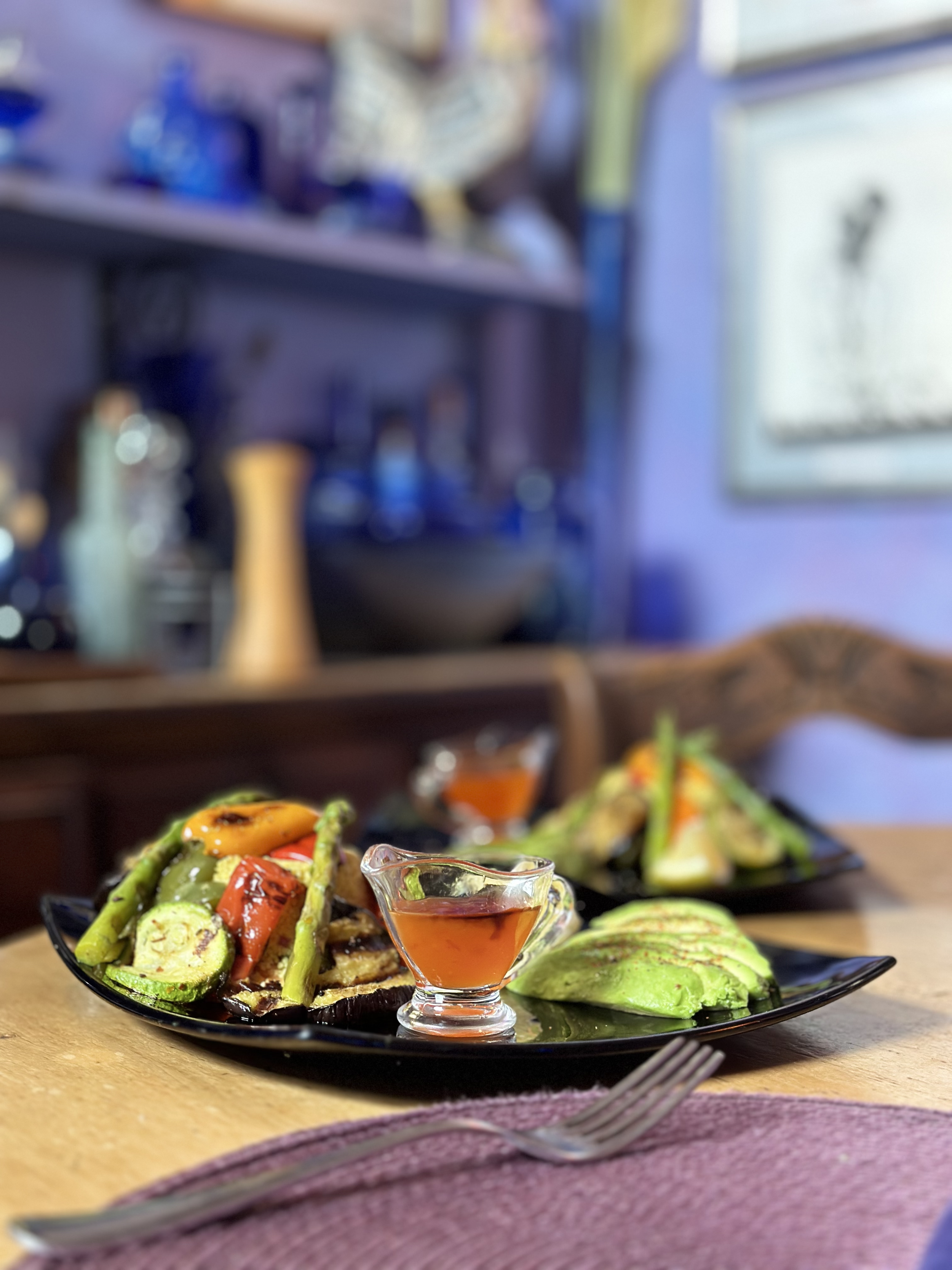 A black plate with grilled vegetables including zucchini, red bell peppers, and asparagus, accompanied by a small glass pitcher of red sauce, on a wooden table with a fork and purple napkin in the foreground, in a restaurant setting.