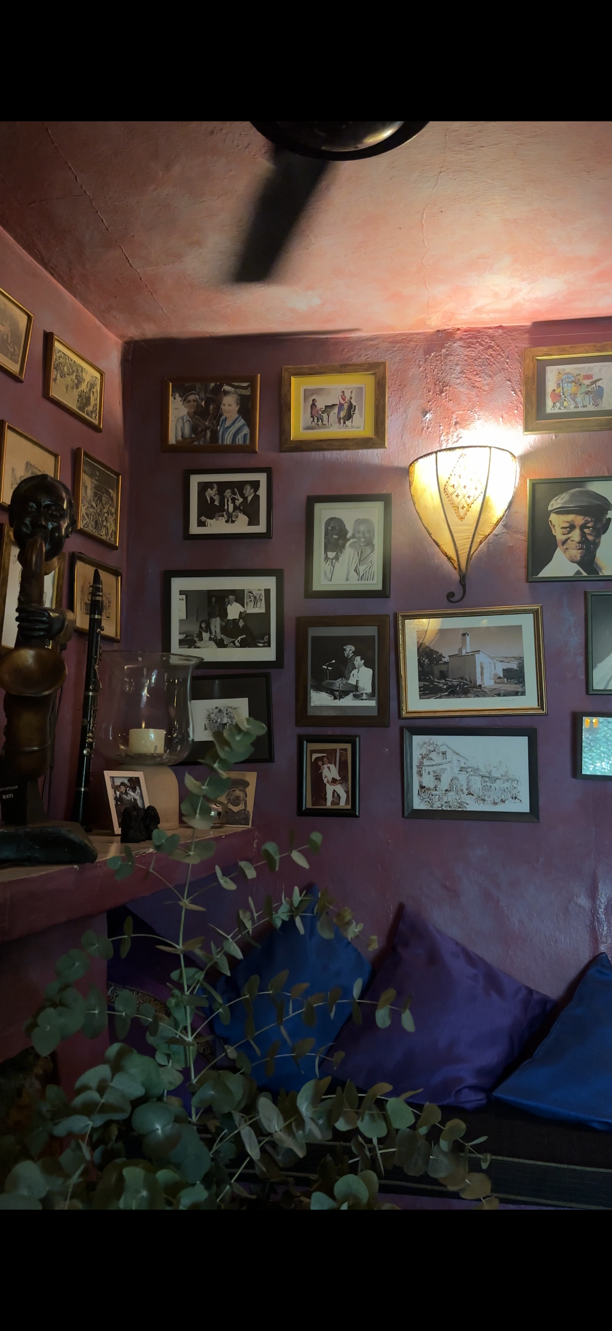 A cozy corner of a room with a pink and purple wall decorated with framed photos and artwork, a wall-mounted light fixture, a table with a candle and plant, and cushions on a bench.