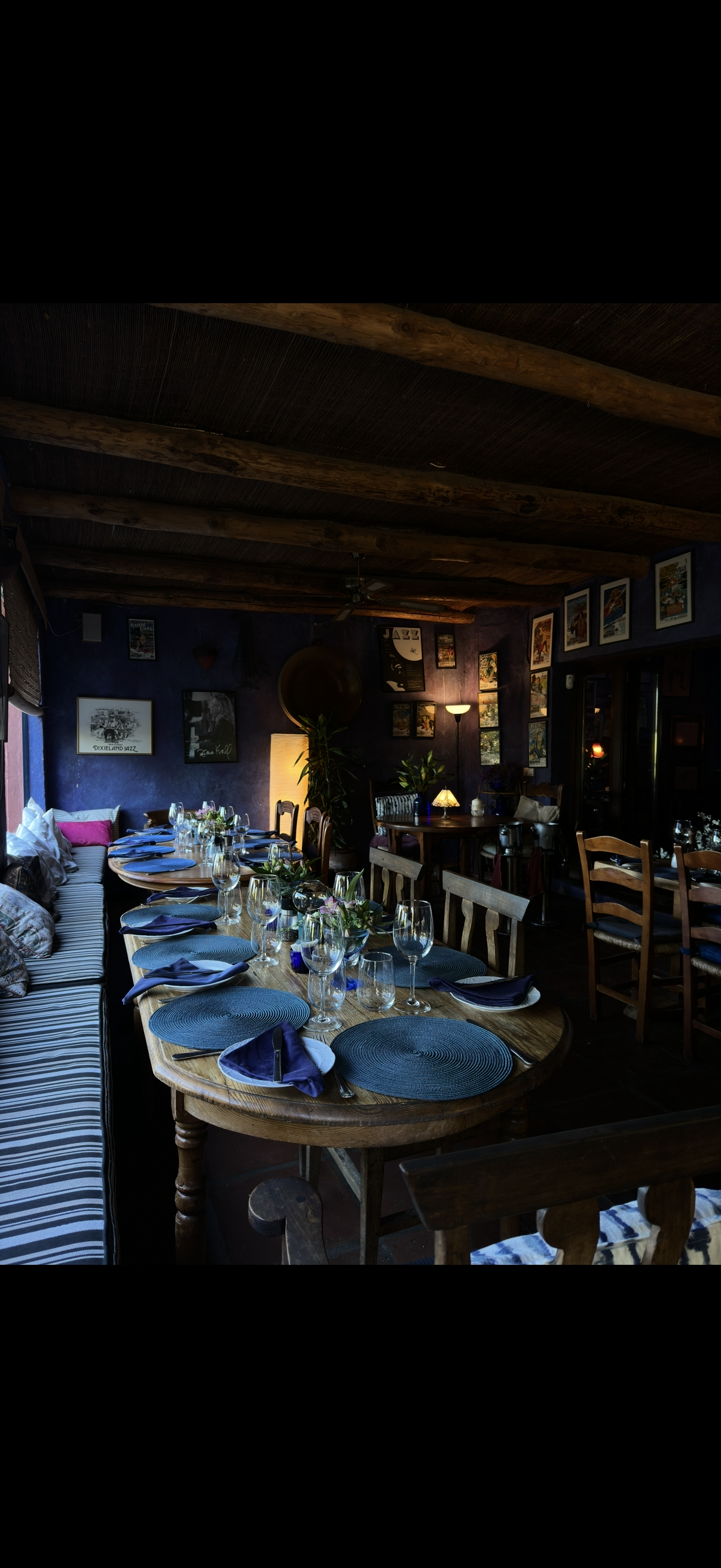 A cozy restaurant interior with a long wooden table set with wine glasses, blue napkins, and placemats. The room has dark-colored walls decorated with framed pictures, and warm lighting from lamps and a floor lamp. Wooden ceiling beams add to the rus