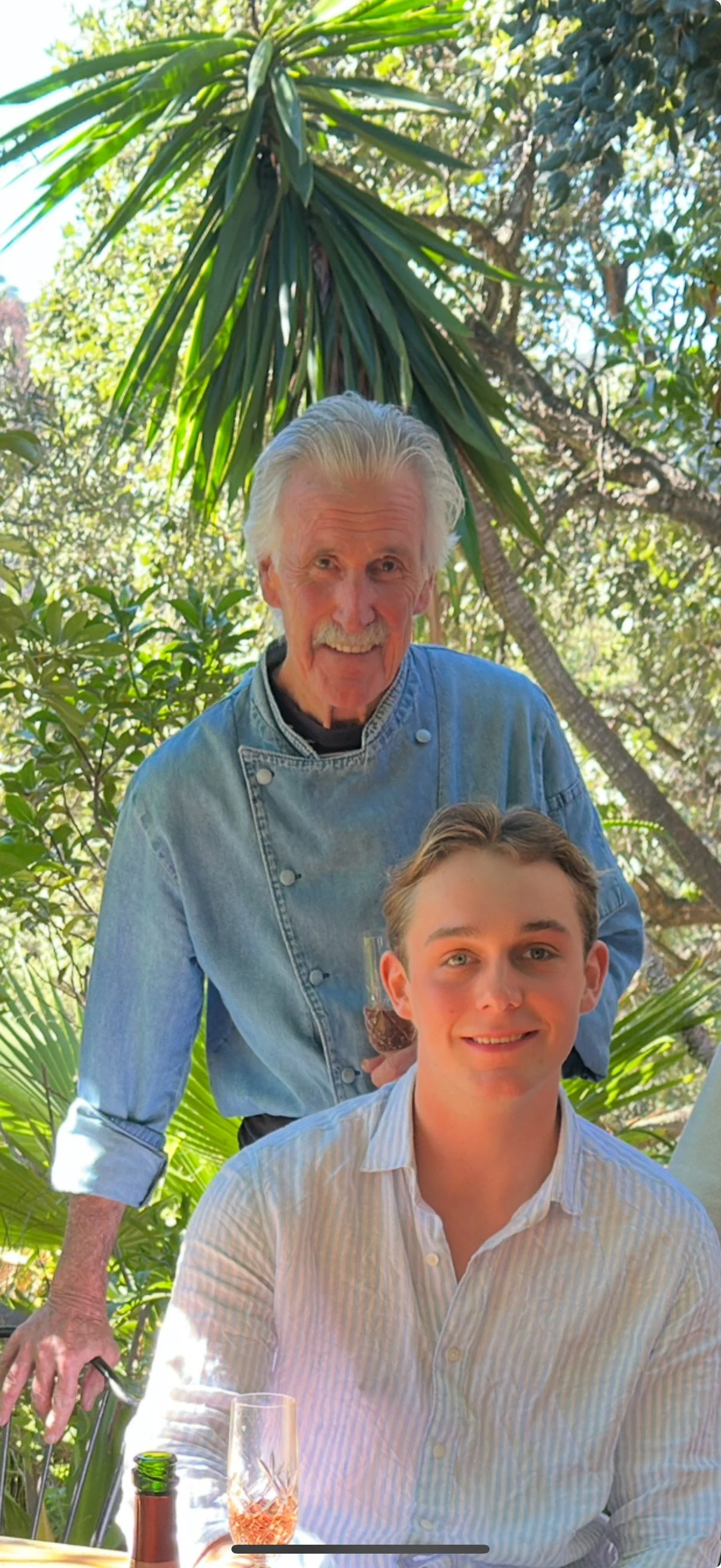 An older man with white hair and mustache wearing a denim chef's coat, standing behind a younger man with brown hair in a white striped shirt, in a lush outdoor setting with trees and sunlight.
