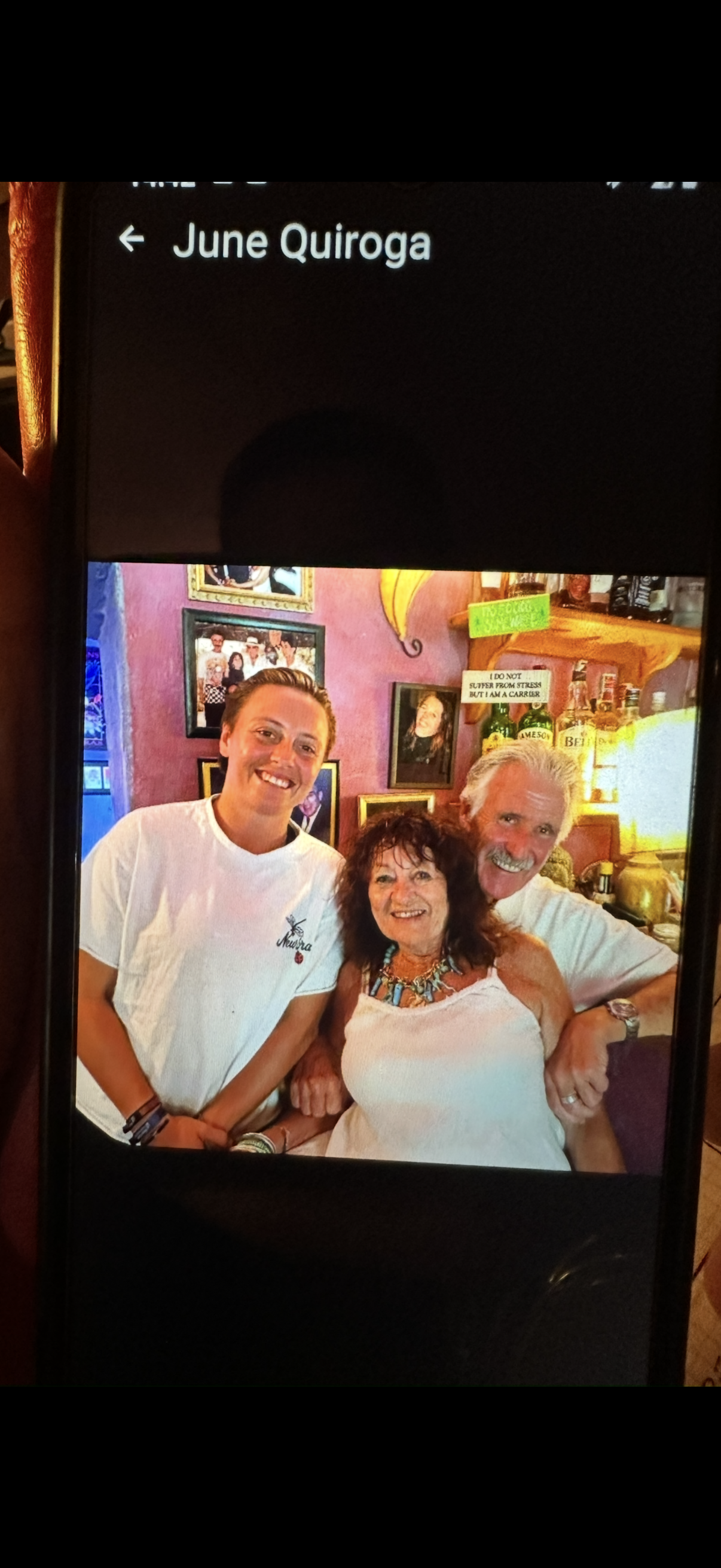 Three smiling people, two women and one man, posing together inside a colorful room with framed pictures and bottles in the background.