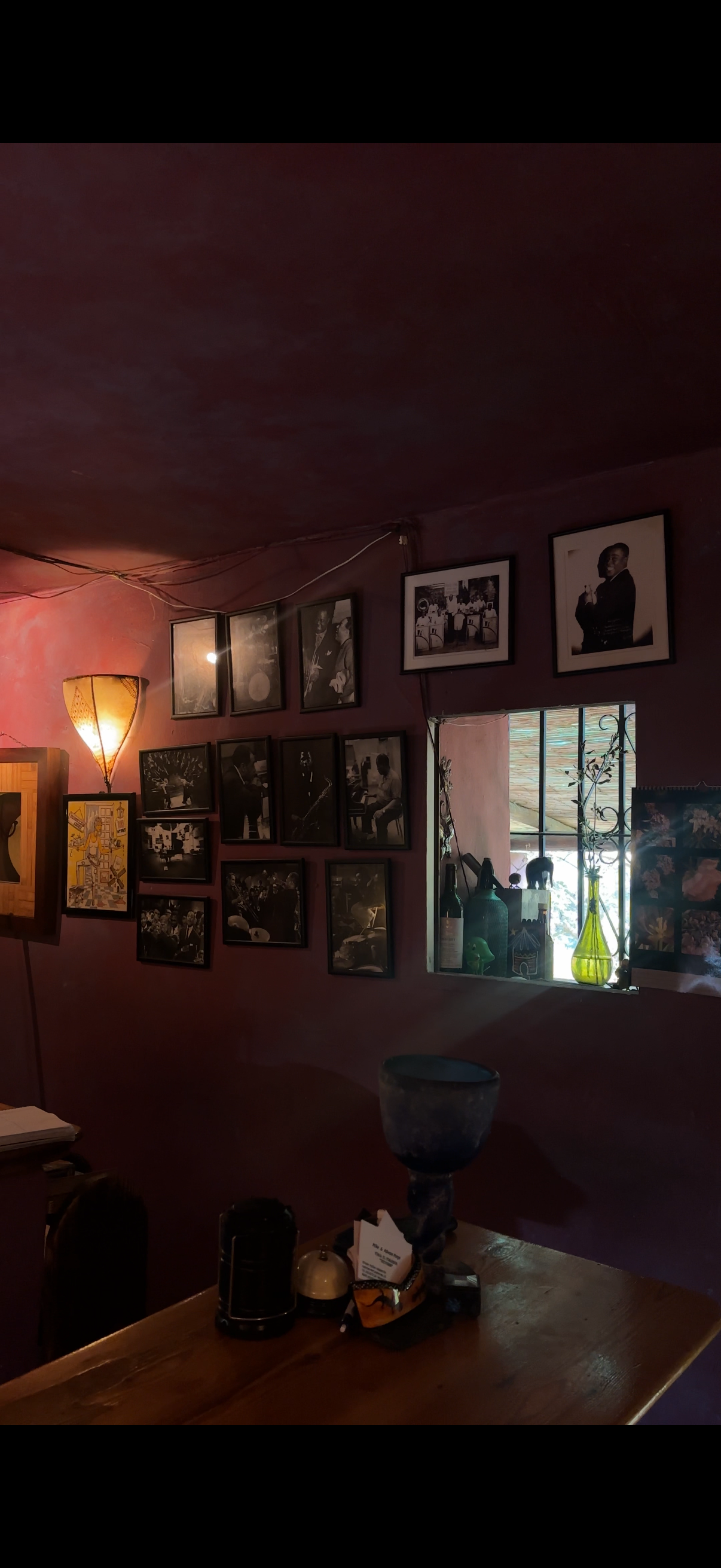 Interior of a cozy room with dark purple walls decorated with black and white photographs, a small window with metal bars, a green vase, wine bottles, and artwork on the windowsill, warm lighting from a floor lamp, and a wooden table with various ite