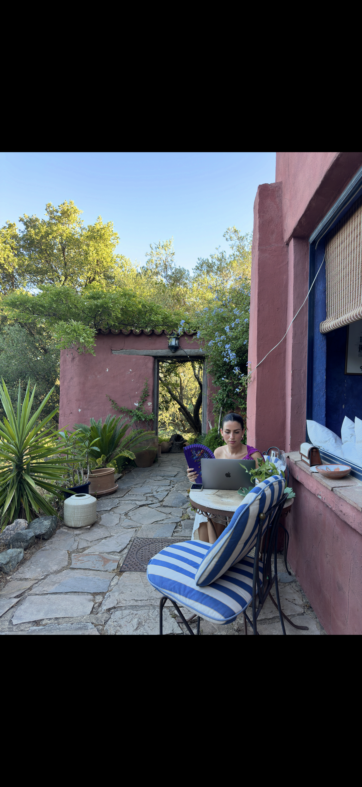 A woman sitting at an outdoor table working on a laptop, holding a purple fan, surrounded by potted plants, against a pink stucco building with a window, in a garden with trees and greenery under a clear blue sky.