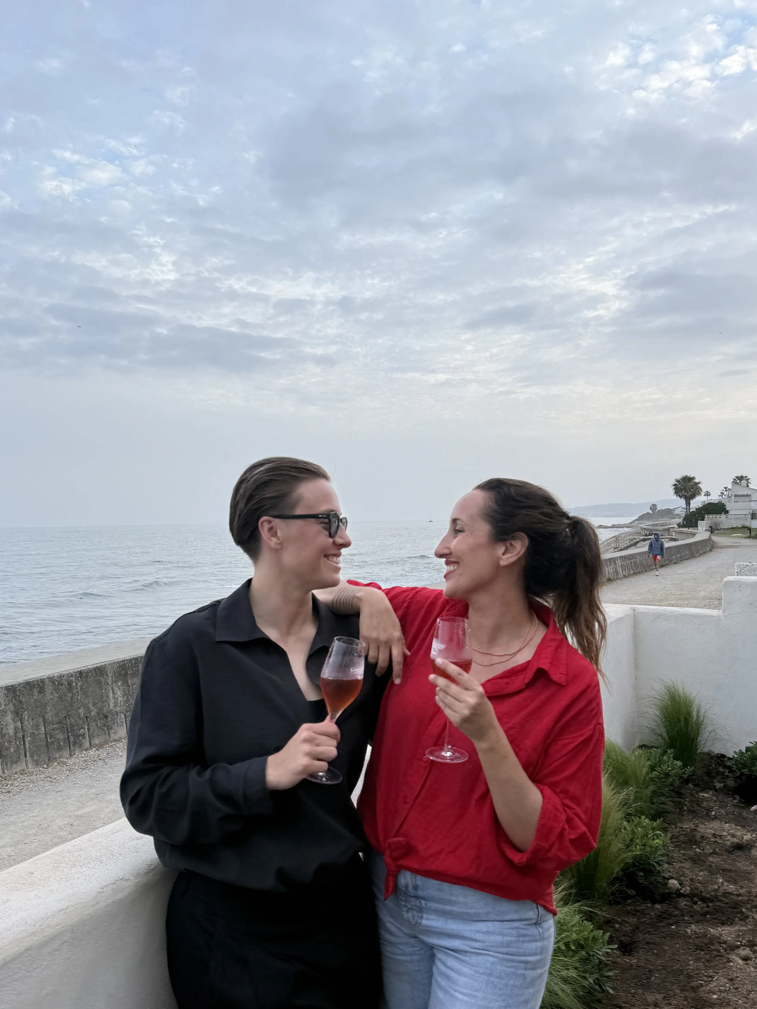 Two women look at each other and smile while holding glasses of rosé wine, standing by a seaside walkway with palm trees and a cloudy sky in the background.