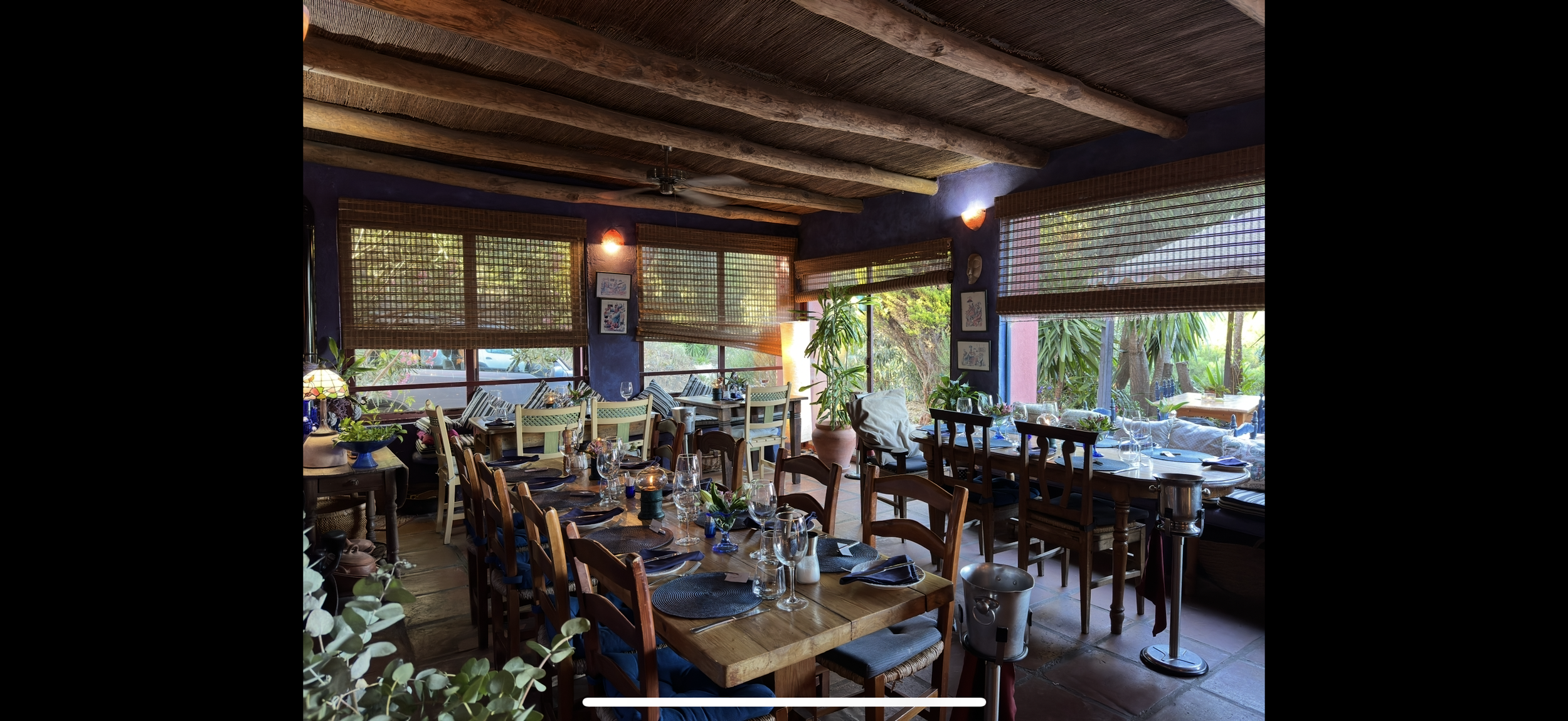 Cozy restaurant interior with wooden beams, large windows with woven shades, and tables set with glassware and napkins, surrounded by plants and colorful artwork.