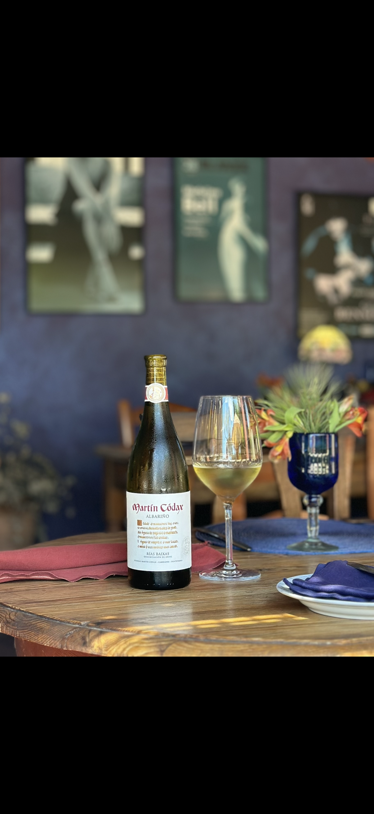 A bottle of white wine, a glass of white wine, and a blue vase with a plant on a wooden table in a cozy restaurant or café setting.
