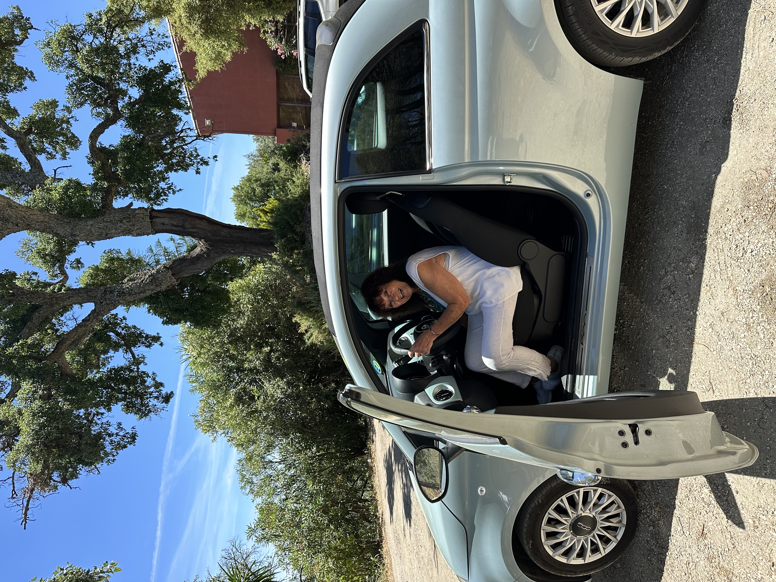 A woman sitting in the driver seat of a silver Fiat 500 car, smiling and looking out the open door, parked under trees with a red building in the background.