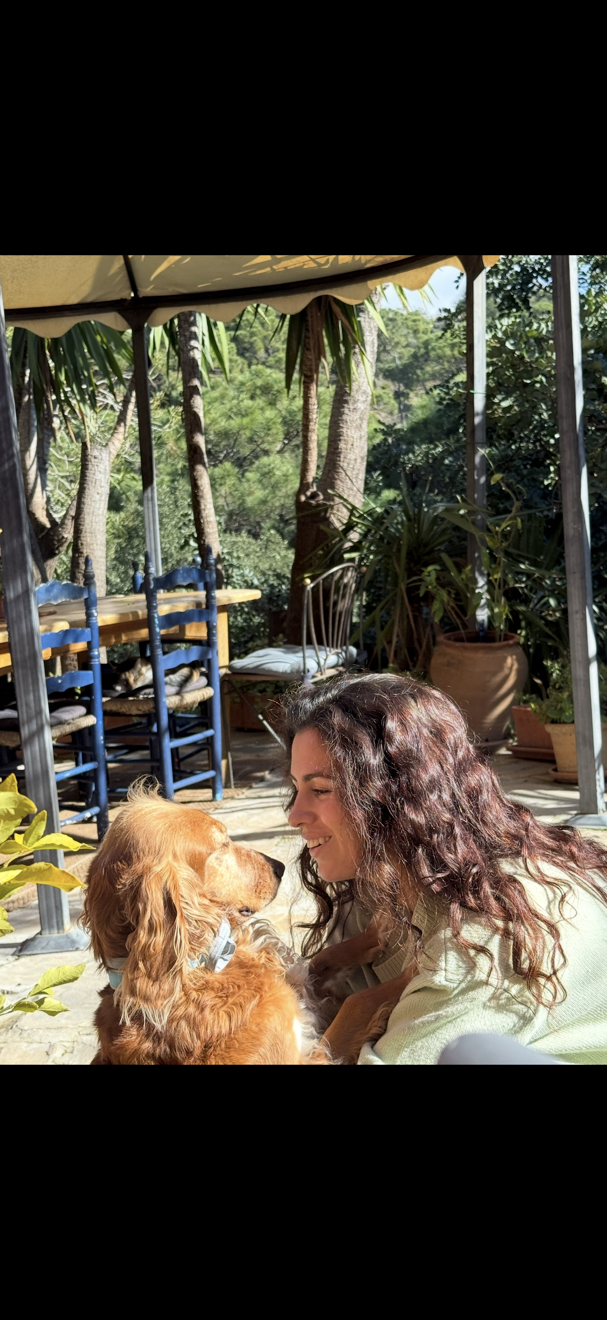 A woman with curly hair smiling at a golden retriever puppy outside on a patio with green plants and trees in the background.