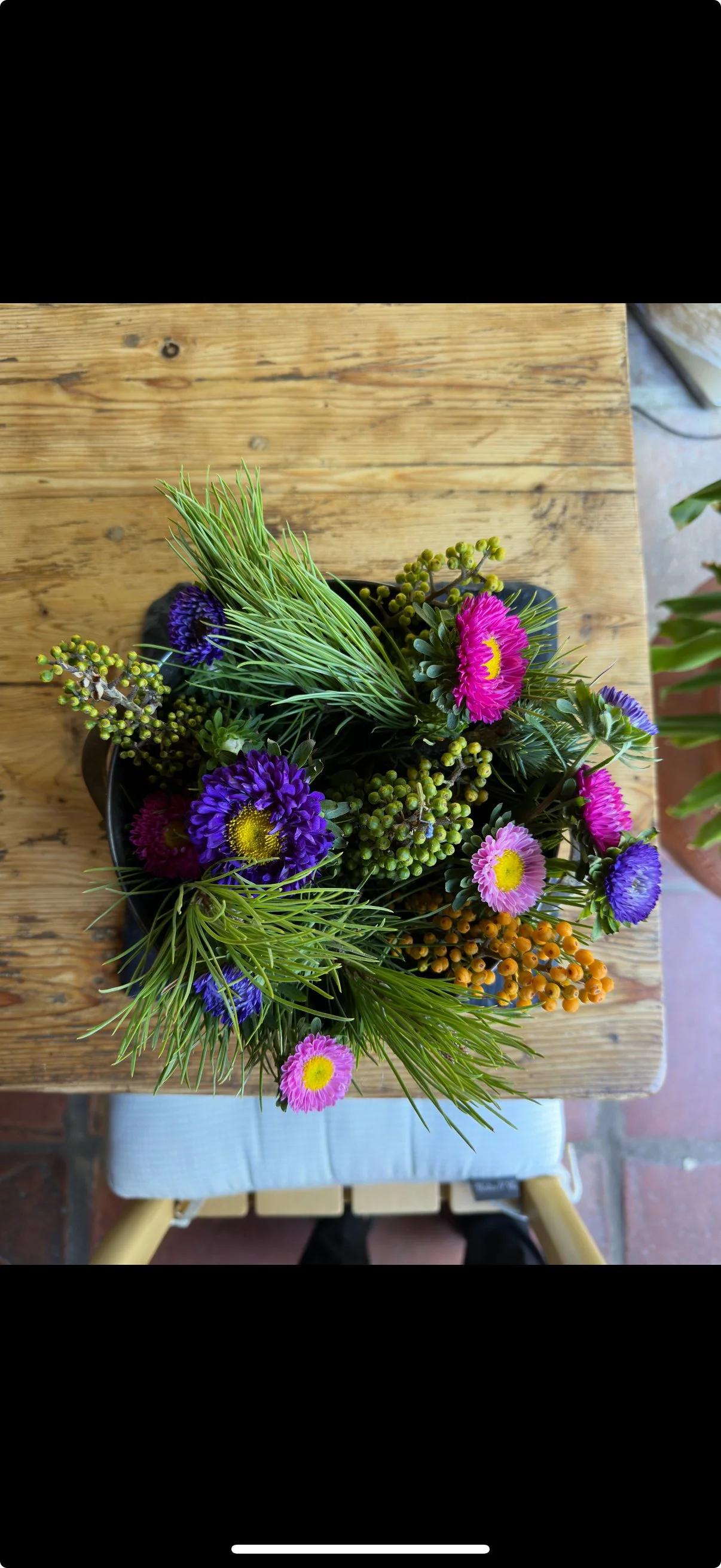 A pot of mixed colorful flowers, including pink and purple daisies, green sprigs, and orange berries, placed on a wooden table.