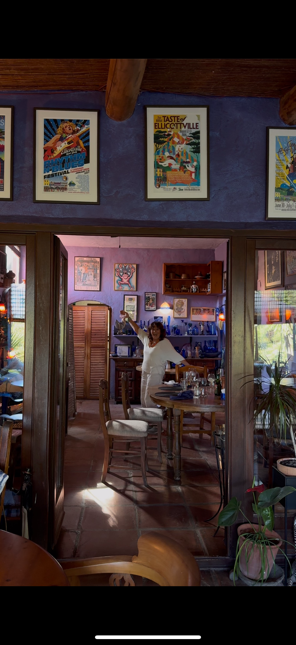 A woman in white clothing standing in a warmly decorated dining room, smiling and raising her arm, with several framed posters hanging on purple walls and a table set with glasses and utensils.