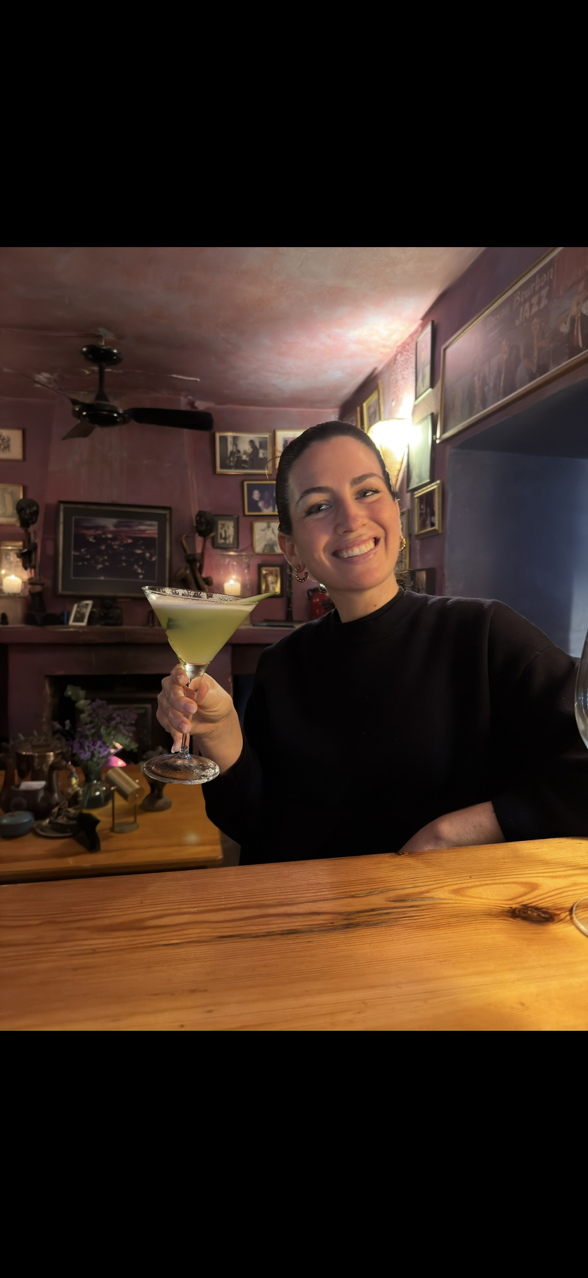 A woman with dark hair smiling and holding a green cocktail in a martini glass inside a dimly lit, vintage-style restaurant with framed pictures on the pinkish wall.