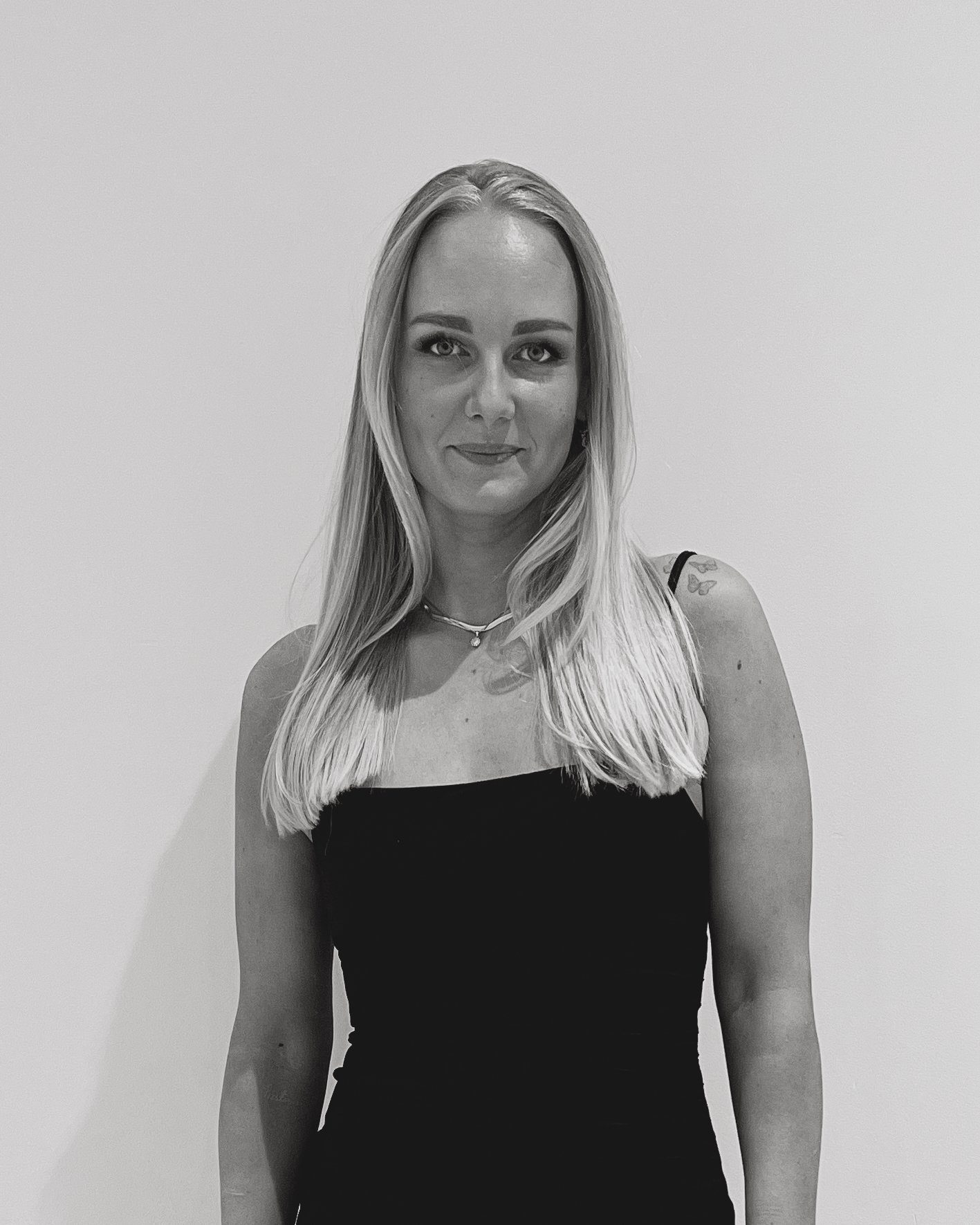 Et svart-hvitt bilde av en kvinne med lang blond hår, iført svart kjole, smilende mot kameraet, stående mot en enkel lys bakgrunn.
