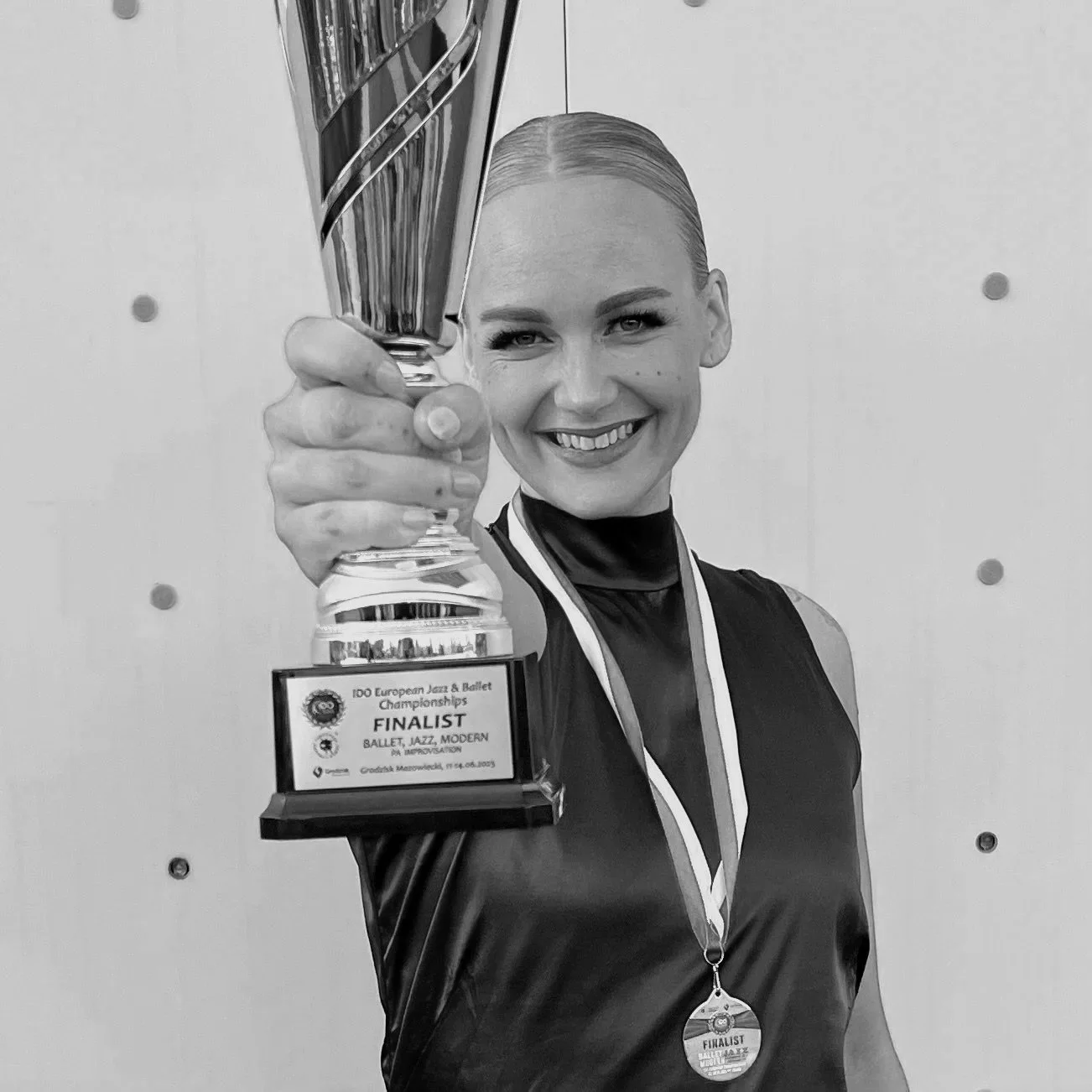 En kvinne smilende og holder opp en pokal som sier finale, finalist, og nevner jazz, ballet og moderne dans, ved en konkurranse.