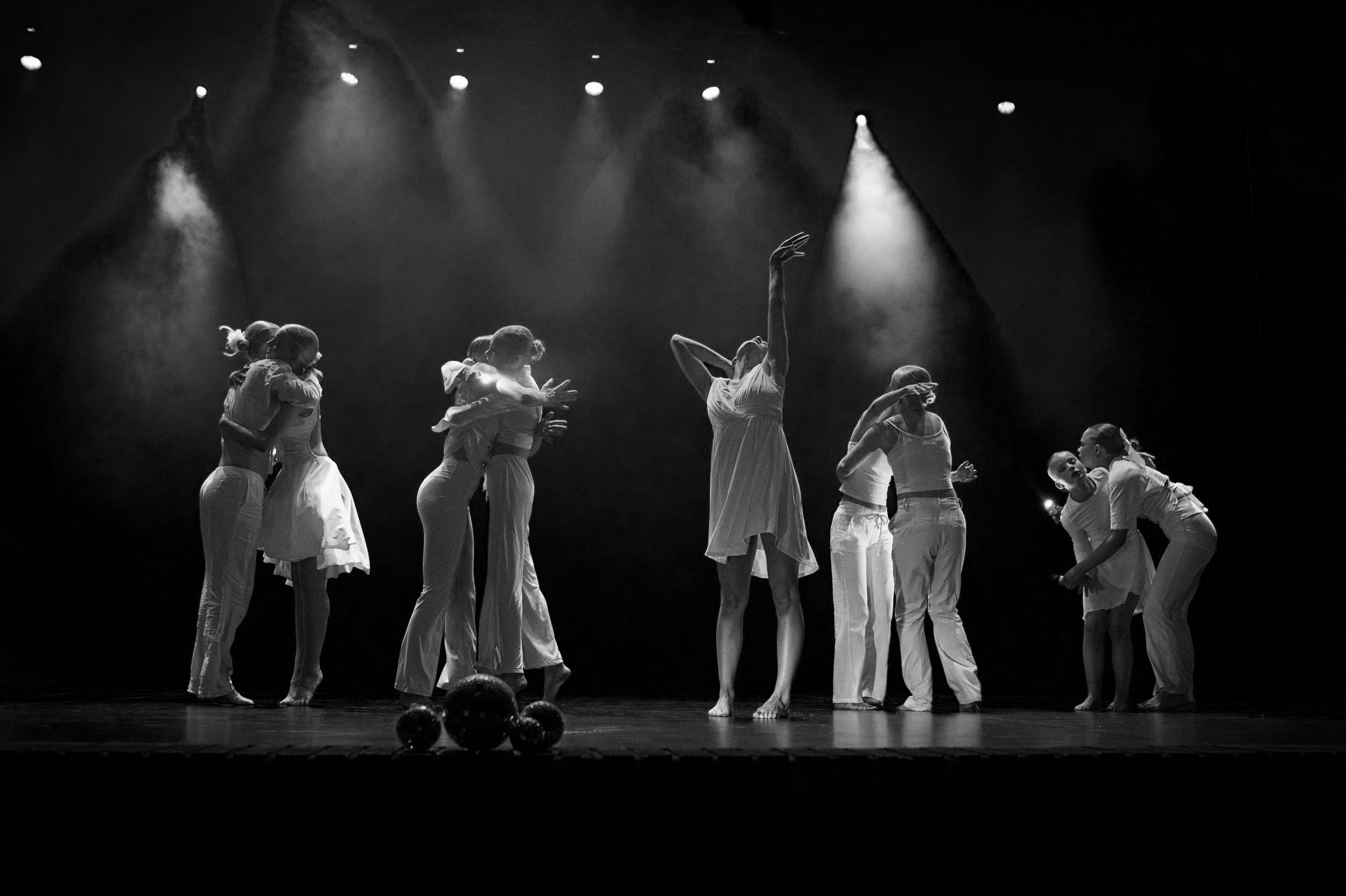 Gruppe av dansere på scenen, kledd i hvite kostymer, under teaterlys, med noen baller på gulvet foran dem.