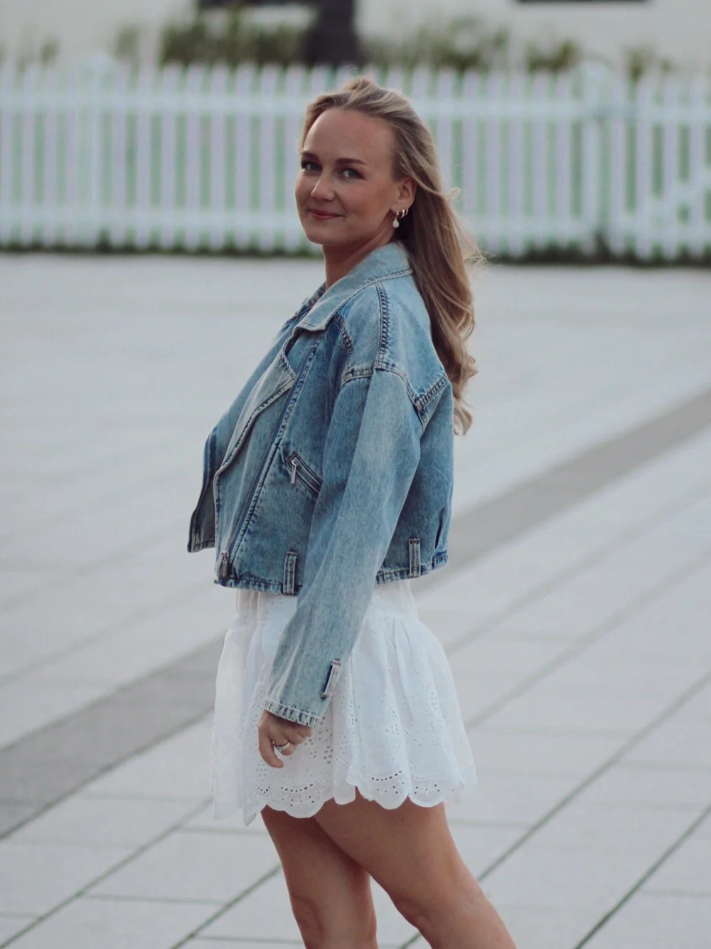 En kvinne med blondt, bølget hår, iført en lys blå denimjakke og en hvit, broderert skjørt står på en utendørs promenade med hvit hekk i bakgrunnen.