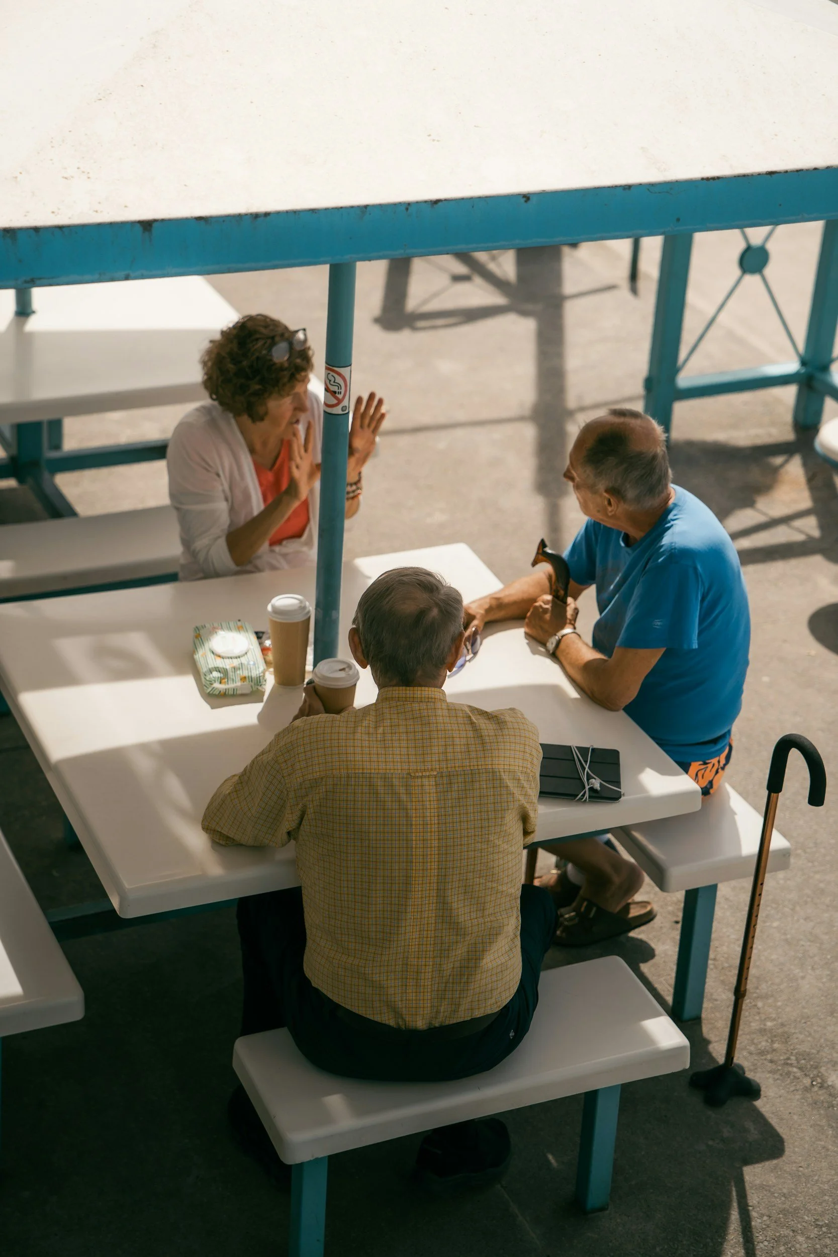 A meeting or discussion taking place at a white outdoor table with four elderly people, two men and two women. One woman appears to be explaining something, while one man listens attentively, and the others are engaged in conversation. Coffee cups and a tissue box are on the table. The setting is outdoors with sunlight and asphalt ground.