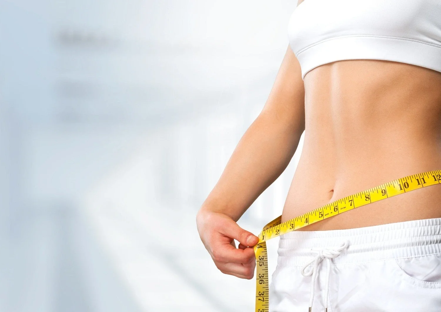 Person measuring their waist with a yellow measuring tape, wearing white shorts and a white top, against a light background.