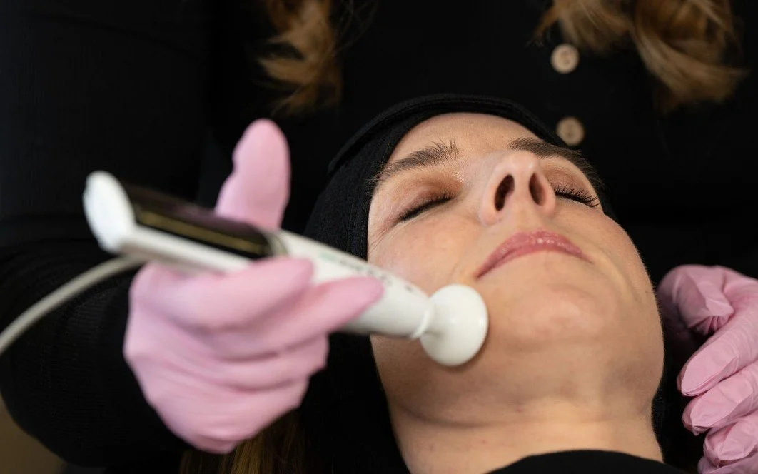A woman receiving a facial treatment with a handheld device on her chin, lying down with her eyes closed, wearing a black headband and pink gloves.