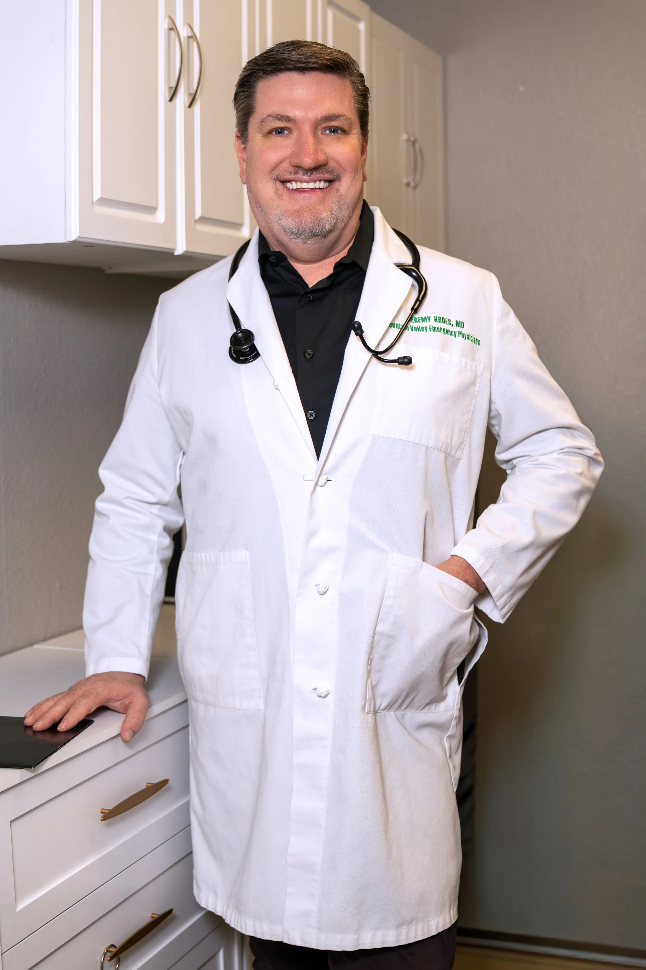 Smiling male doctor with short dark hair, wearing a white coat and stethoscope around his neck, standing in a medical office or clinic, with cabinets in the background.