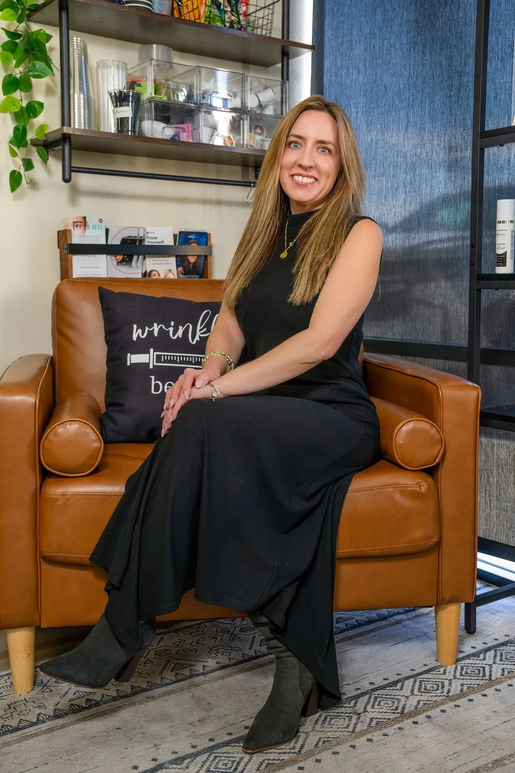 A woman sitting on a brown leather couch with a black pillow that has white text and a syringe graphic. She has long, light brown hair, and is wearing a black sleeveless dress, jewelry, and gray boots. Behind her, there are shelves with various office supplies, papers, and informational brochures. The setting appears to be an office or waiting area with a modern style.
