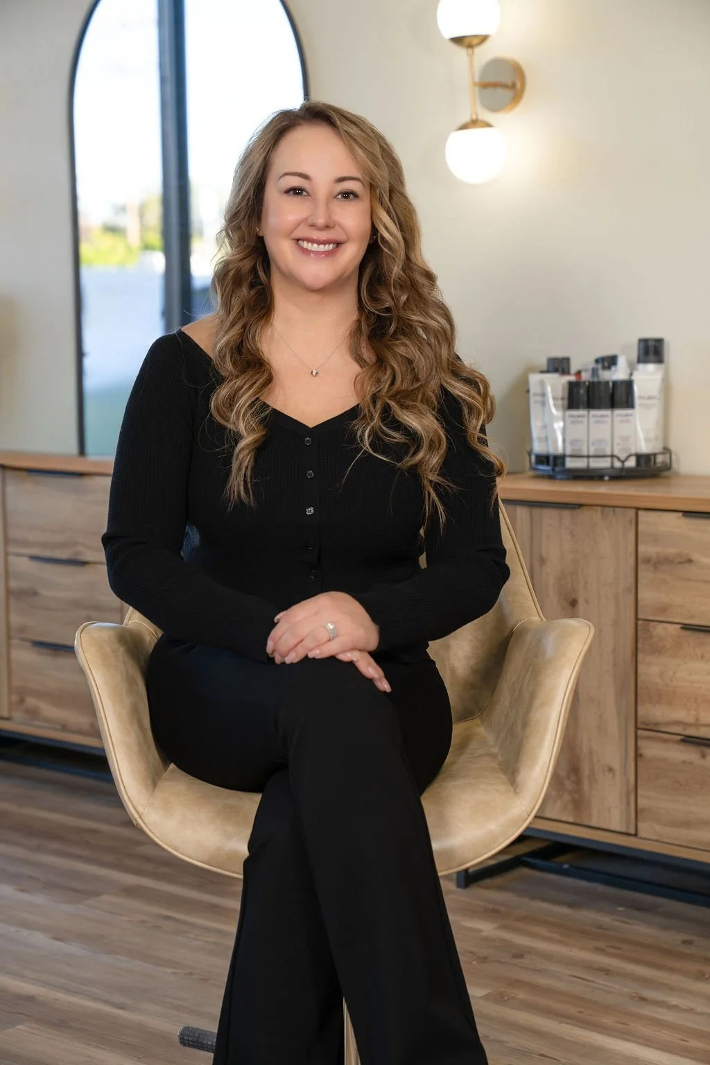 A smiling woman with long, wavy brown hair sitting on a beige chair in a modern room with a large arched window, wooden furniture, and a wall-mounted light fixture.