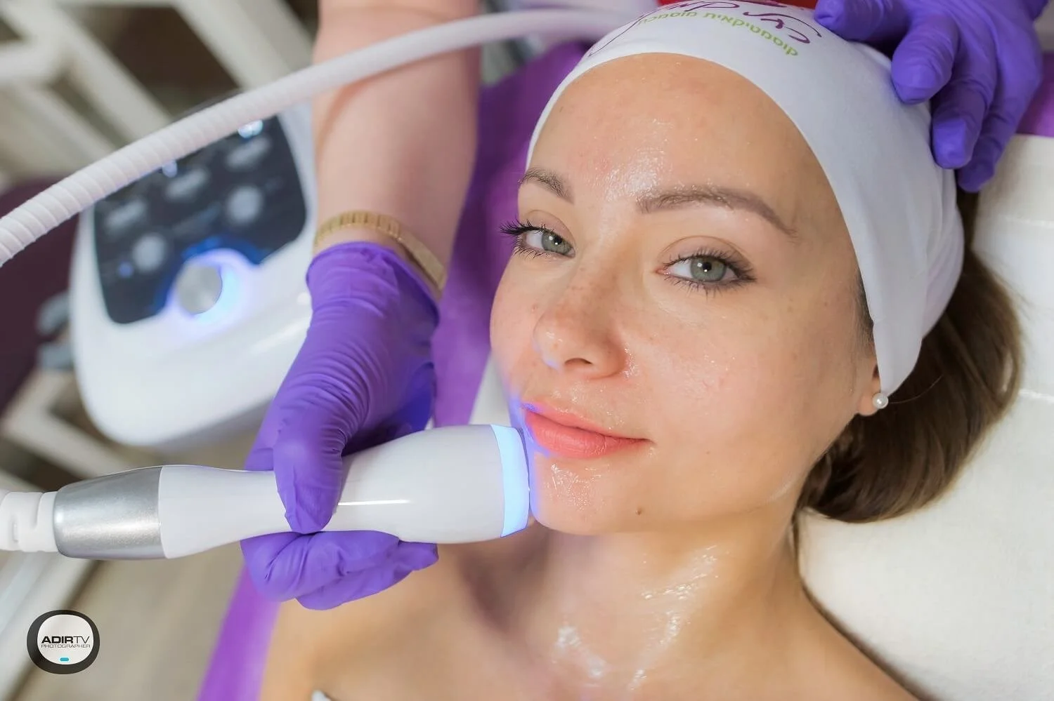 A woman with clear skin and light makeup receives a facial treatment with a handheld device emitting blue light, while lying down with her head on a pillow, wearing a white headband, and a person wearing purple gloves performs the procedure.