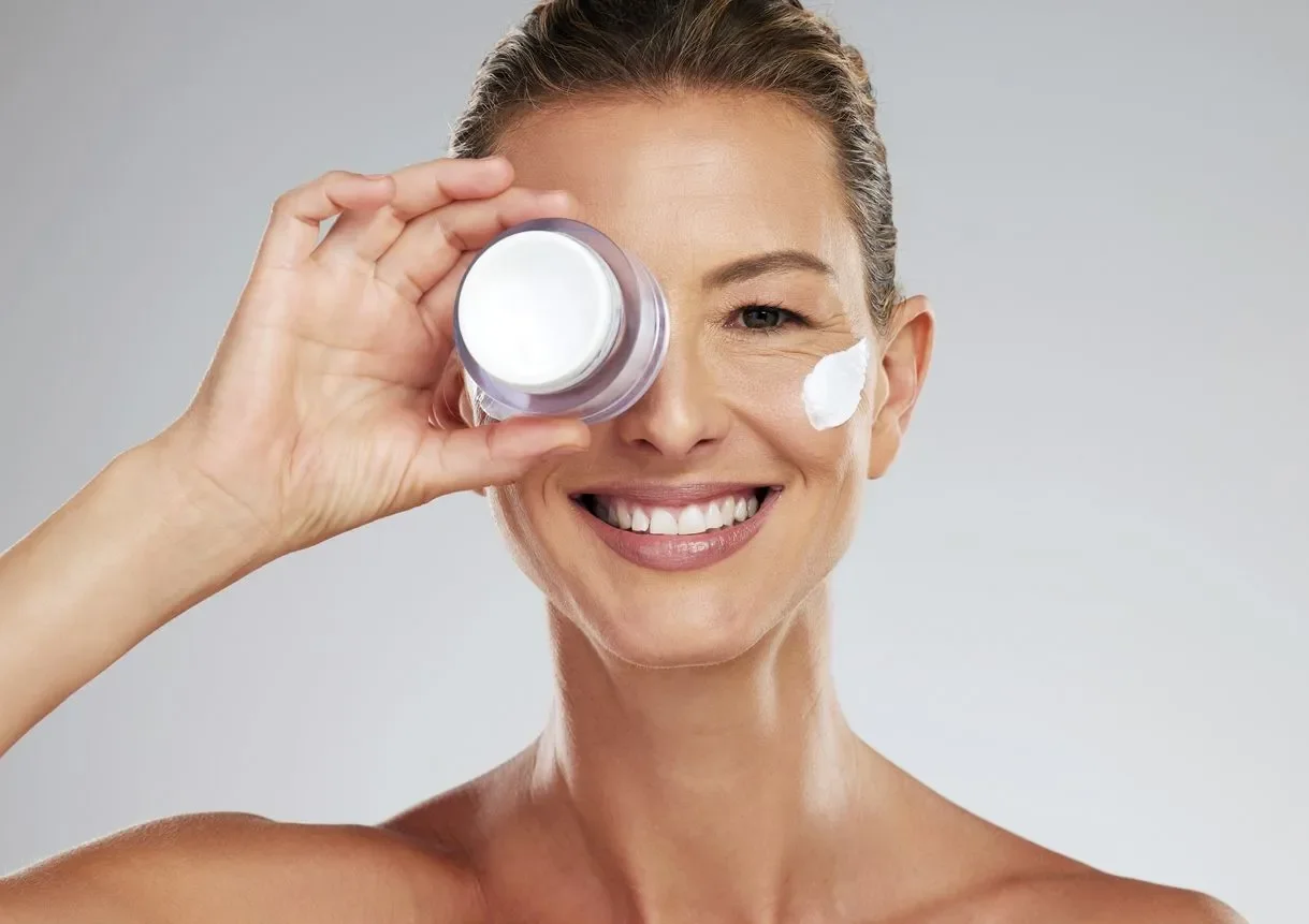A woman with a broad smile applying skin cream to her face, holding a jar of cream near her eye, against a plain background.