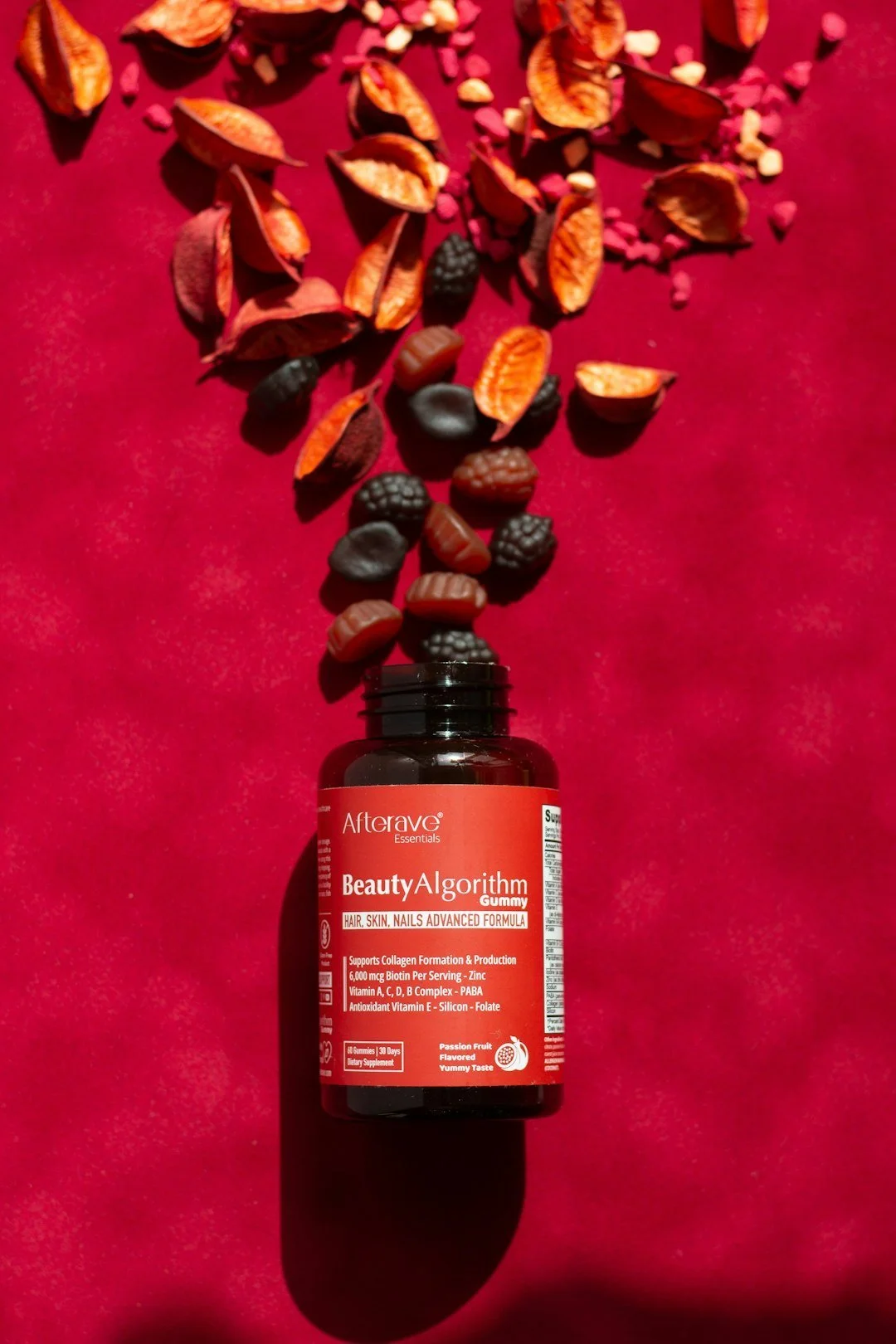 A bottle of Afterave Beauty Algorithm Gummy supplement on a red surface with assorted gummy candies spilling out of the bottle, including blackberries and strawberries.