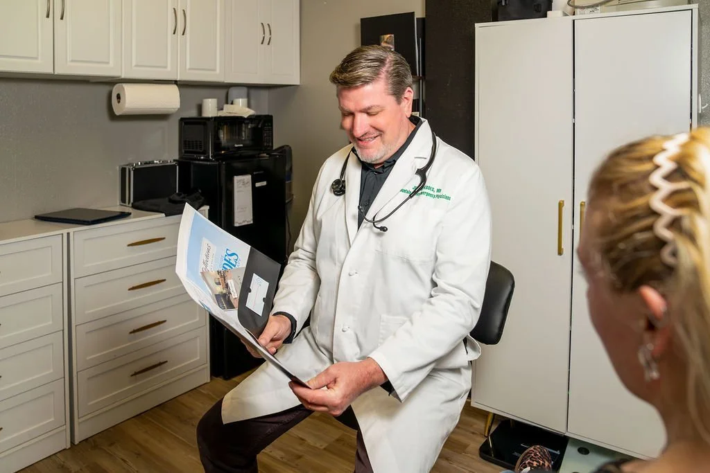 A male doctor in a white coat and stethoscope sitting and reading a medical magazine in a room with cabinets and kitchen appliances.