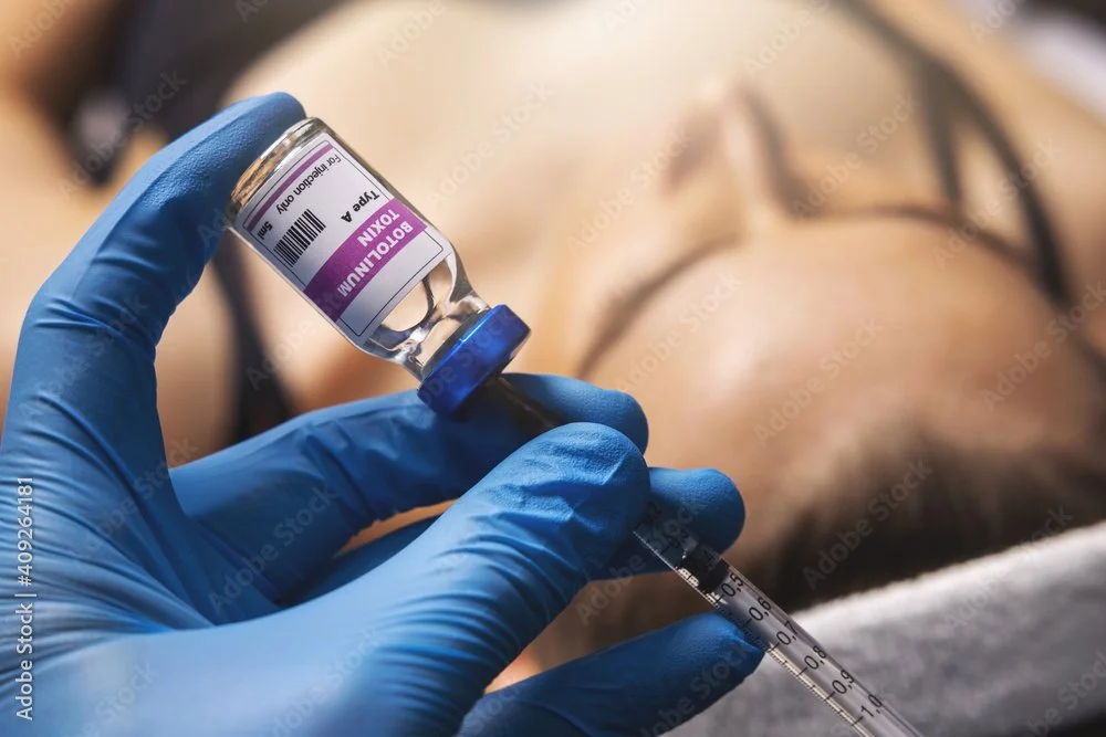 A healthcare worker wearing blue gloves holding a vial of the COVID-19 vaccine and a syringe, about to administer the vaccine to a person lying down.