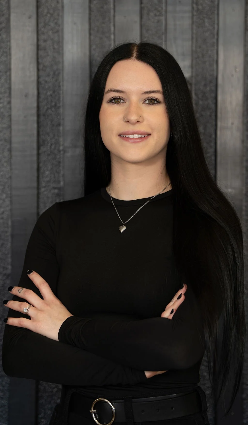 A young woman with long black hair, wearing a black long sleeve top, a heart-shaped necklace, and rings, standing in front of a wooden wall.