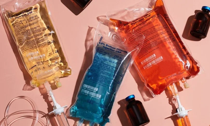 Three IV fluid bags on a pink surface, filled with yellow, blue, and orange liquids, with black medication vials nearby.