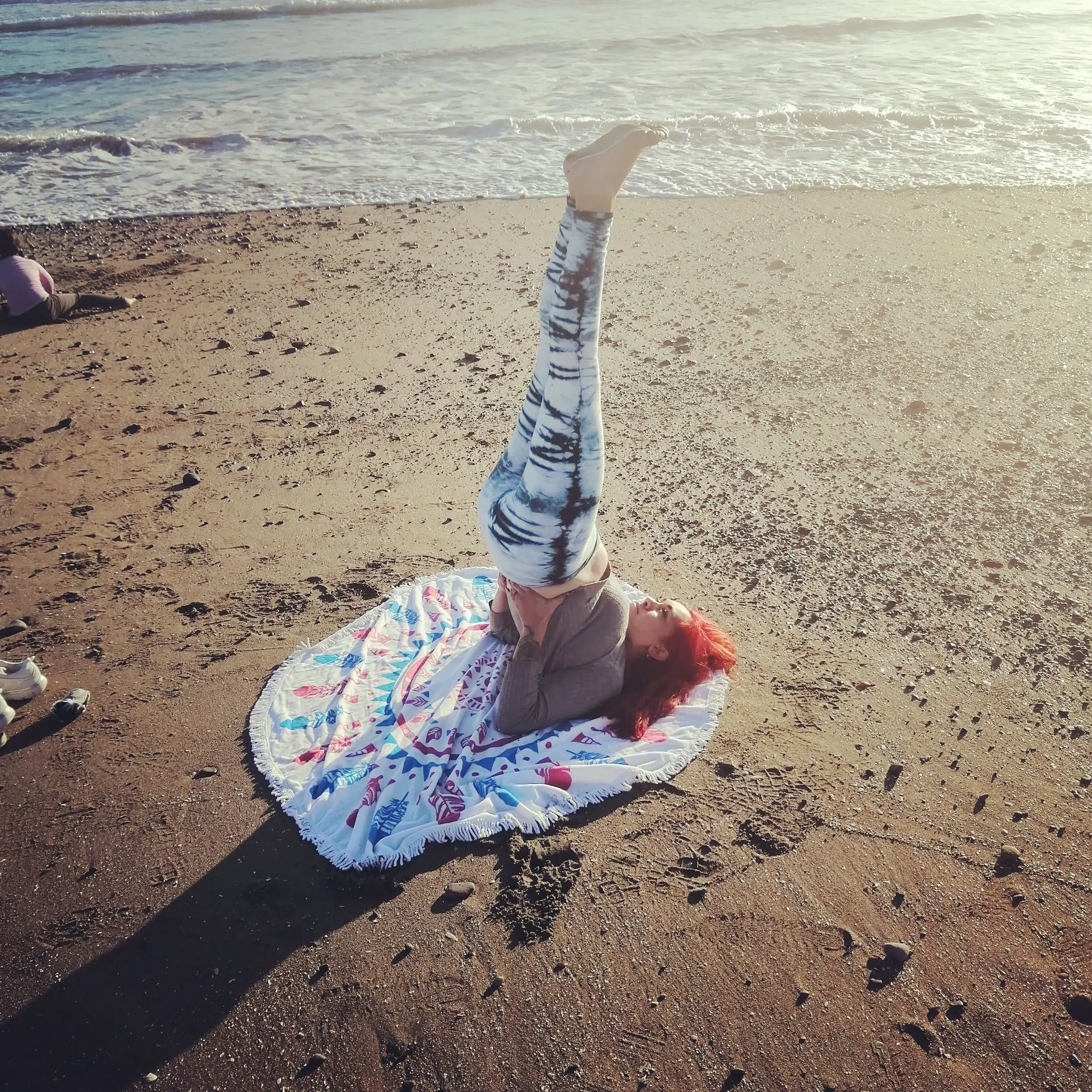 Niña con cabello rojizo haciendo una voltereta hacia atrás en la playa, sobre una toalla con estampado colorido, cerca del mar, con el sol brillante y olas en el fondo.