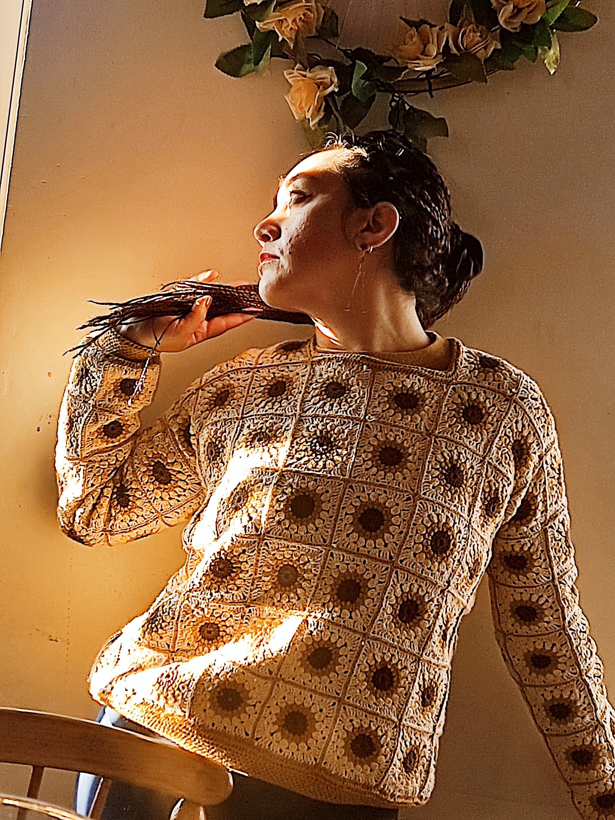 Retrato de una mujer con cabello trenzado, usando una blusa con diseño floral, en un ambiente con luz cálida y plantas colgantes en el techo, tocando su cuello con la mano izquierda.