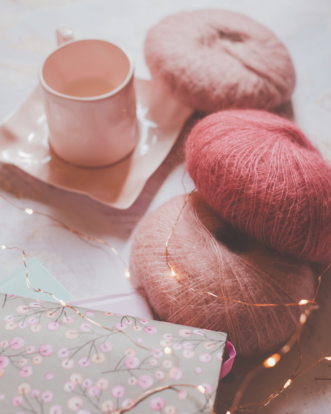 Tres ovillos de lana en tonos rosados y una taza rosa en una bandeja, decorados con luces de hadas y una caja con diseño floral.