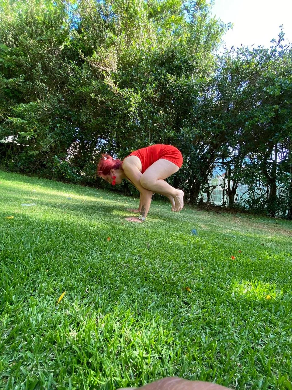 Mujer con cabello rojo en posición de equilibrio en una pierna en un césped verde, rodeada de arbustos y árboles, vestida con ropa roja.