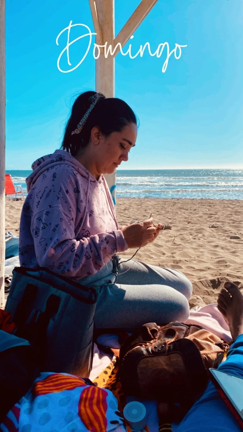 Joven mujer sentada en la playa bajo una estructura de madera, vestida con sudadera de color lila y pantalones claros, mirando su teléfono móvil, con arena y mar al fondo.