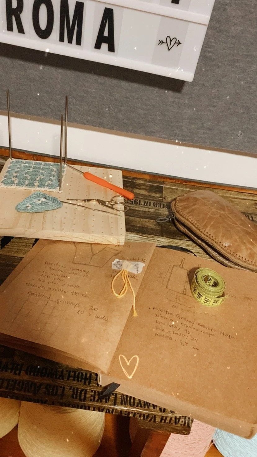 Mesa de trabajo con notas y mediciones, una bolsa de cuero, tijeras, filamentos y cinta métrica. En el fondo, un cartel que dice 'ROMA' con un corazón y flechas.