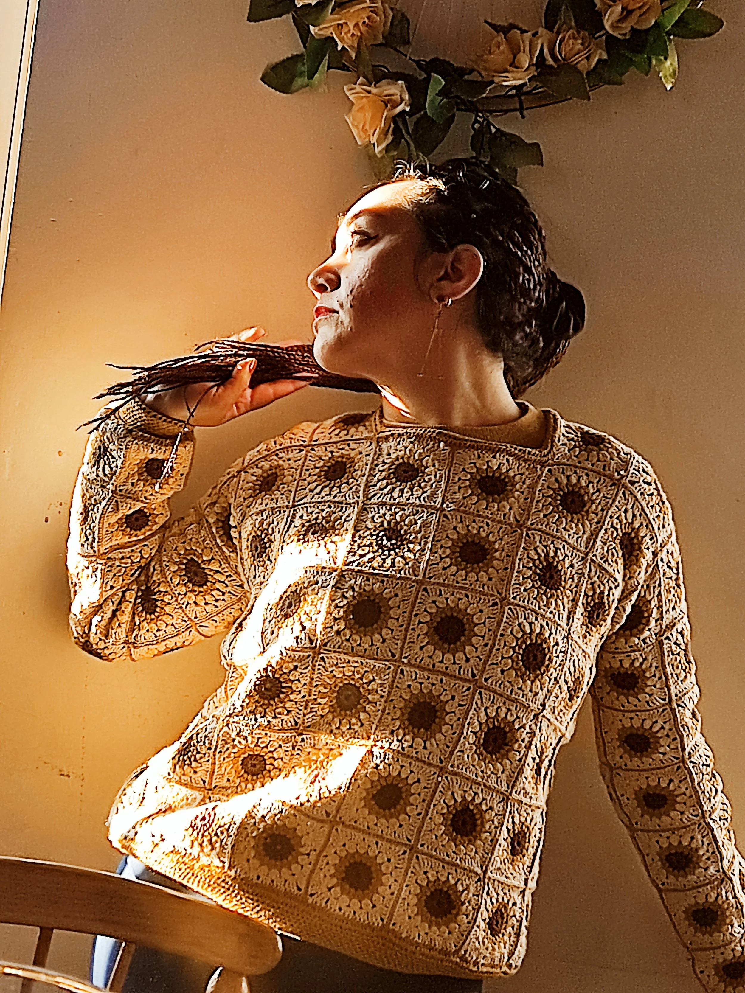 Una mujer con trenzas en el cabello, usa un suéter de patrones florales y círculos, y está en un espacio con una lámpara y una mesa de madera. Está tocando su cuello y tiene un pendiente largo en la oreja izquierda.