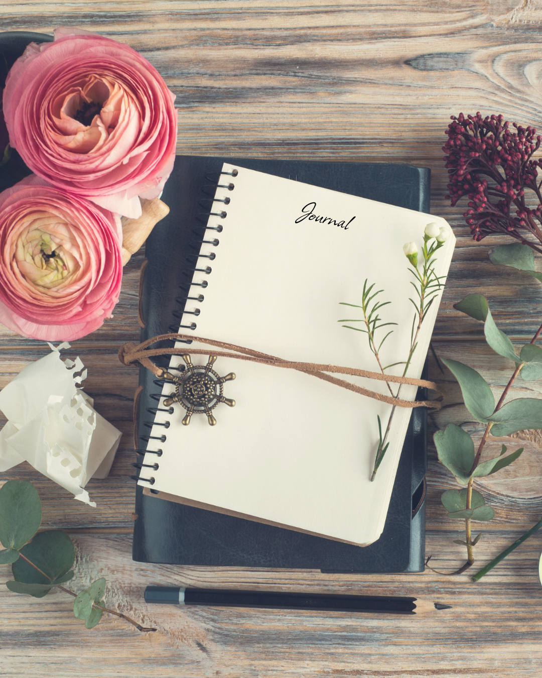 Un cuaderno con la palabra 'Journal' en la portada, rodeado de flores y una pluma en una mesa de madera.