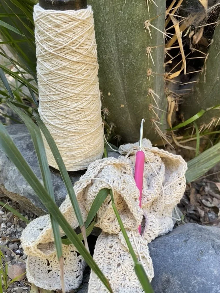 Un ovillo de hilo de color crema y varias piezas de ganchillo hechas a mano, acompañadas de una aguja de ganchillo rosada, en un entorno natural con plantas y rocas.