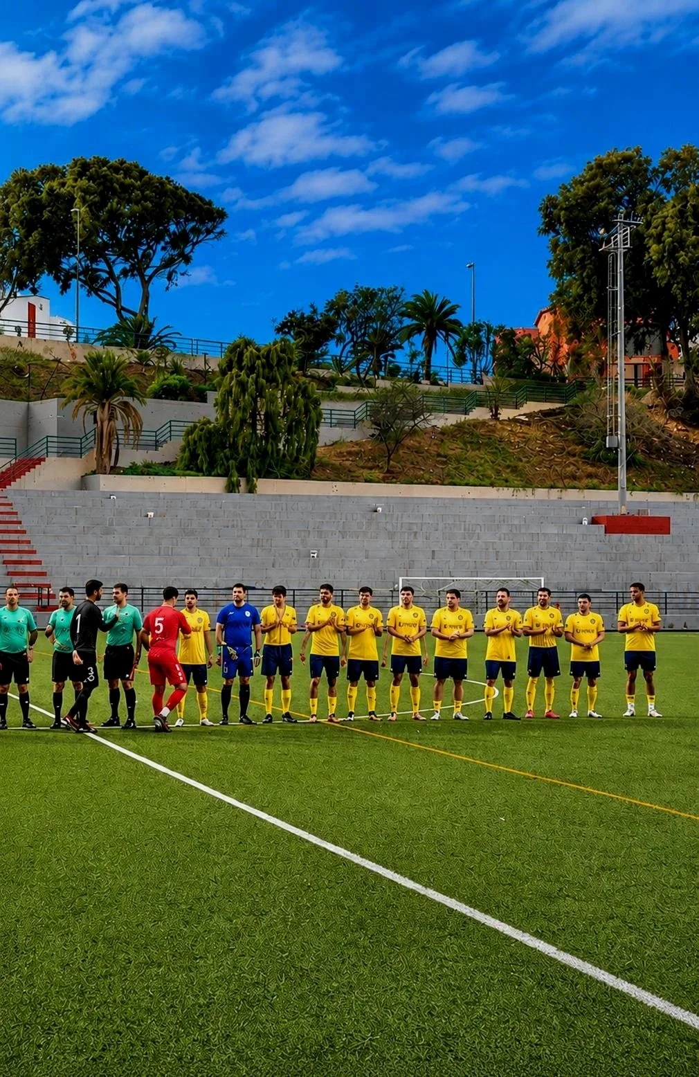 A soccer team in yellow jerseys lined up on a green field, with two goalkeepers in different uniforms, and a referee in a turquoise shirt, standing near the sideline of the field with a stadium and trees in the background.