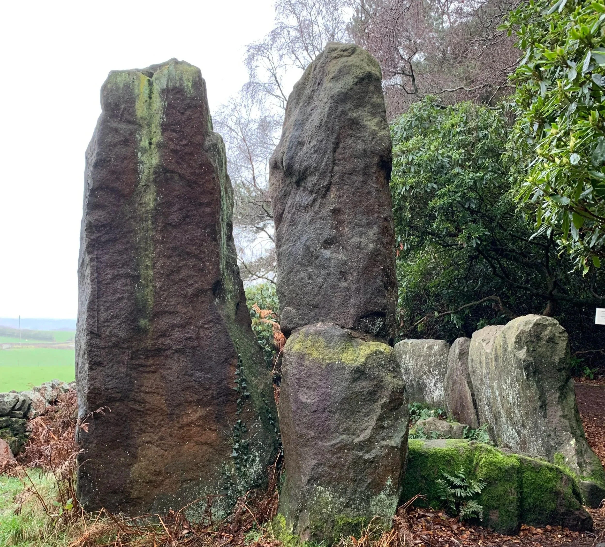 A row of large standing and fallen stones in a garden or park surrounded by green foliage and trees.