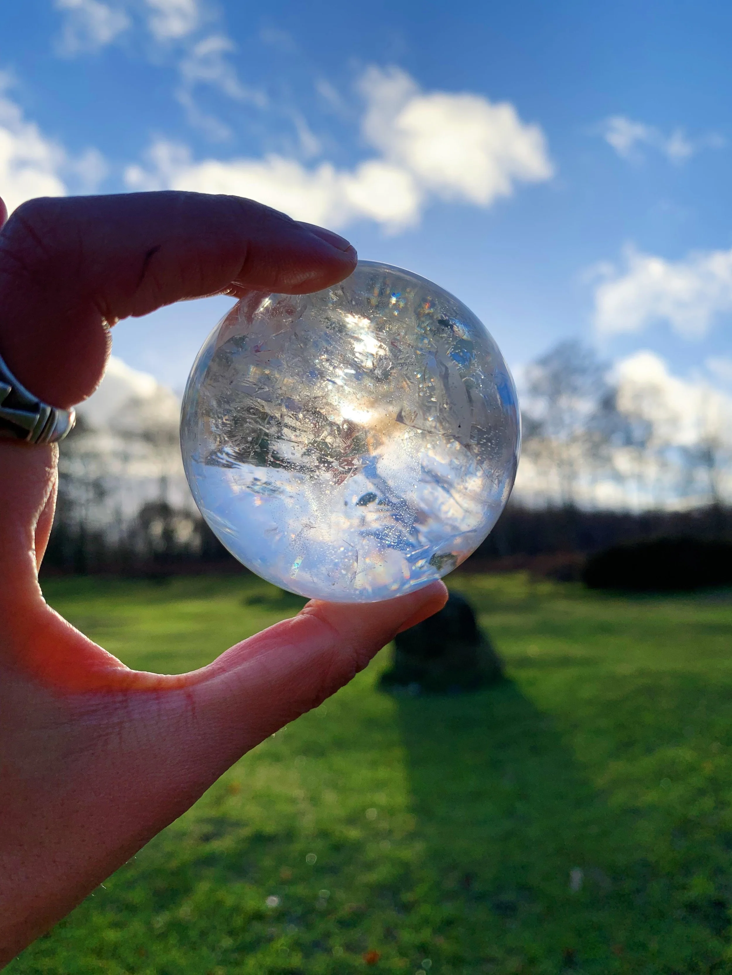 Person holding a clear glass sphere outdoors with a green grassy area and trees in the background, under a blue sky with white clouds.