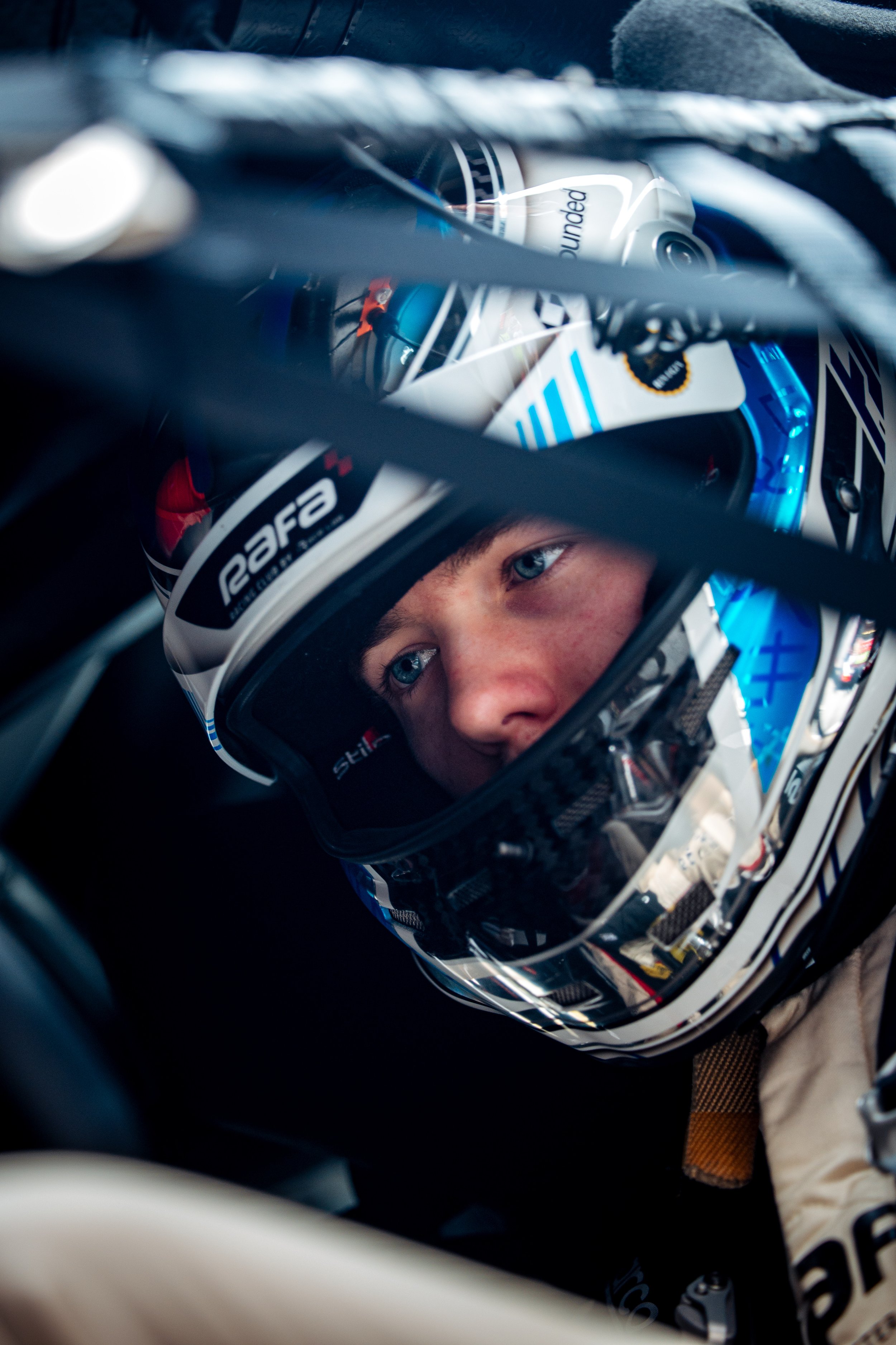 Close-up of a race car driver wearing a racing helmet with a focused expression, sitting inside a vehicle.