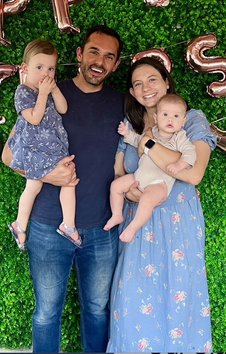 A family of four celebrating a birthday in front of a green leafy backdrop with pink balloon letters spelling out 'Love'. The father is holding a young girl in a blue floral dress, and the mother is holding a baby dressed in beige. Both the father and mother are smiling at the camera.