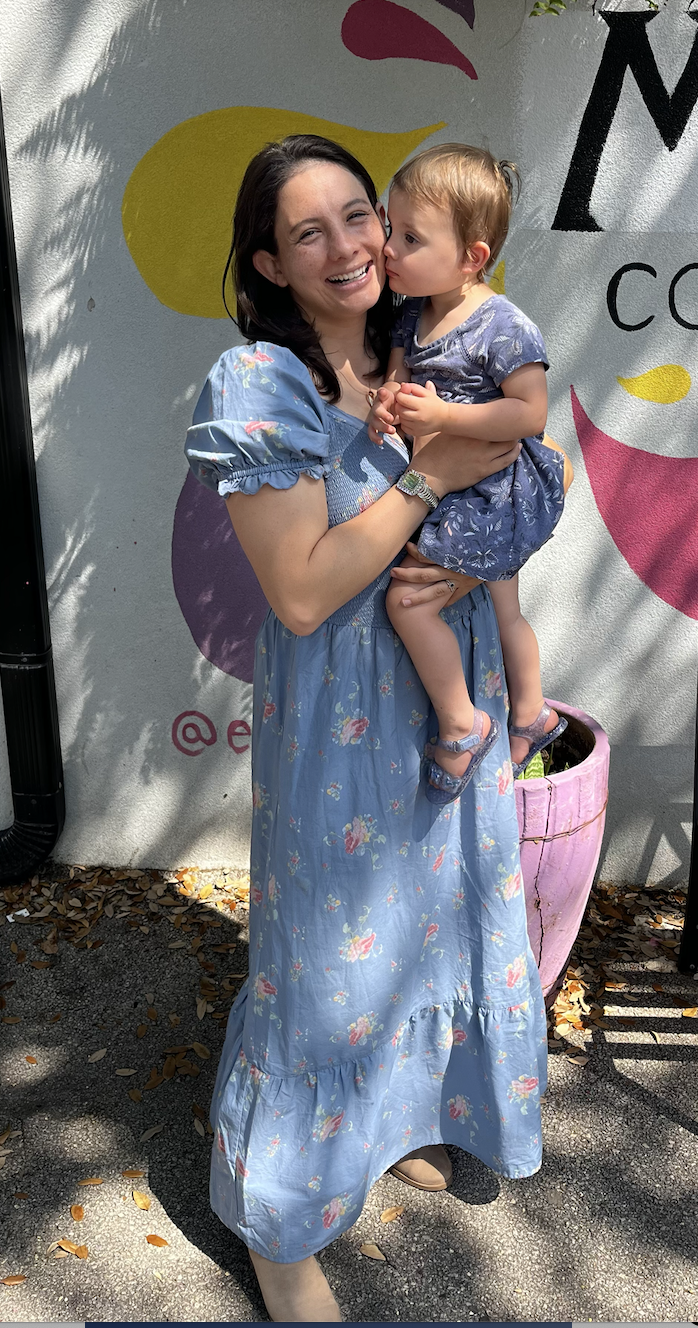 A woman in a floral blue maxi dress holding a young girl in a similar dress against a colorful painted wall with a pink planter and fallen leaves on the ground.