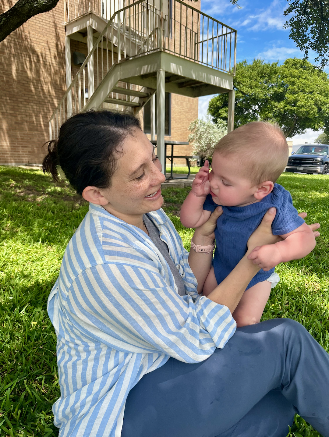 A woman with short dark hair and freckles smiling while holding a small blond toddler in a park with green grass, trees, and a brick building in the background.