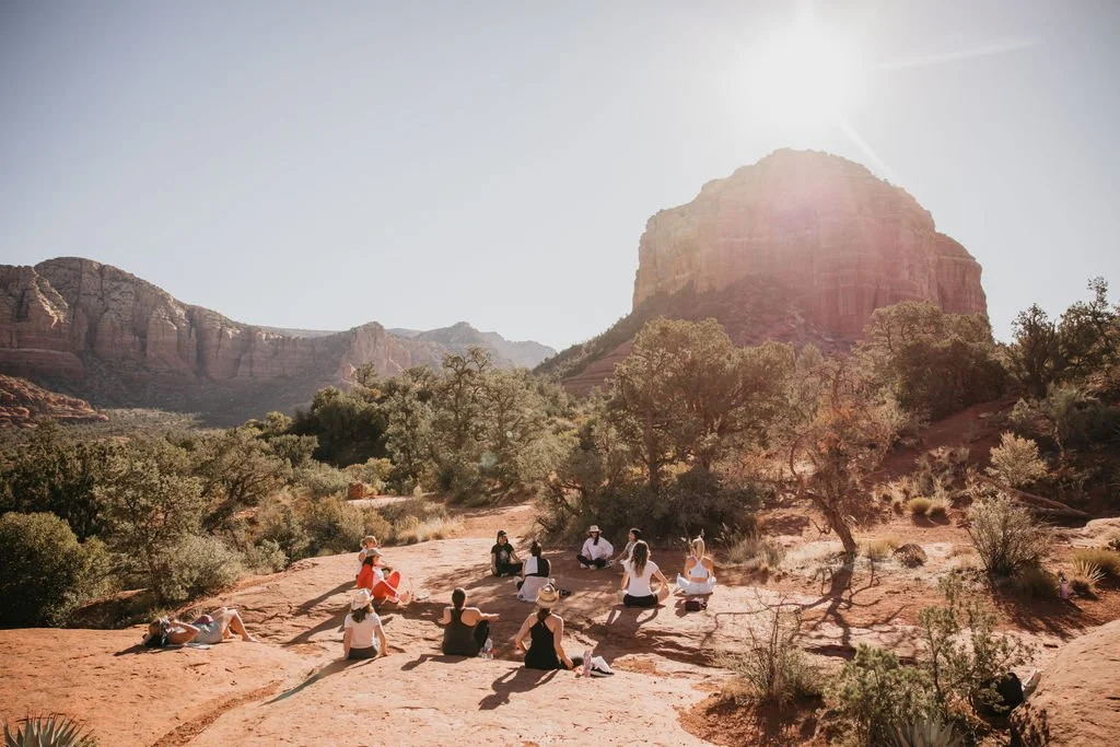 People sitting on a rocky desert landscape surrounded by trees and large rock formations, with sunlight shining from above.