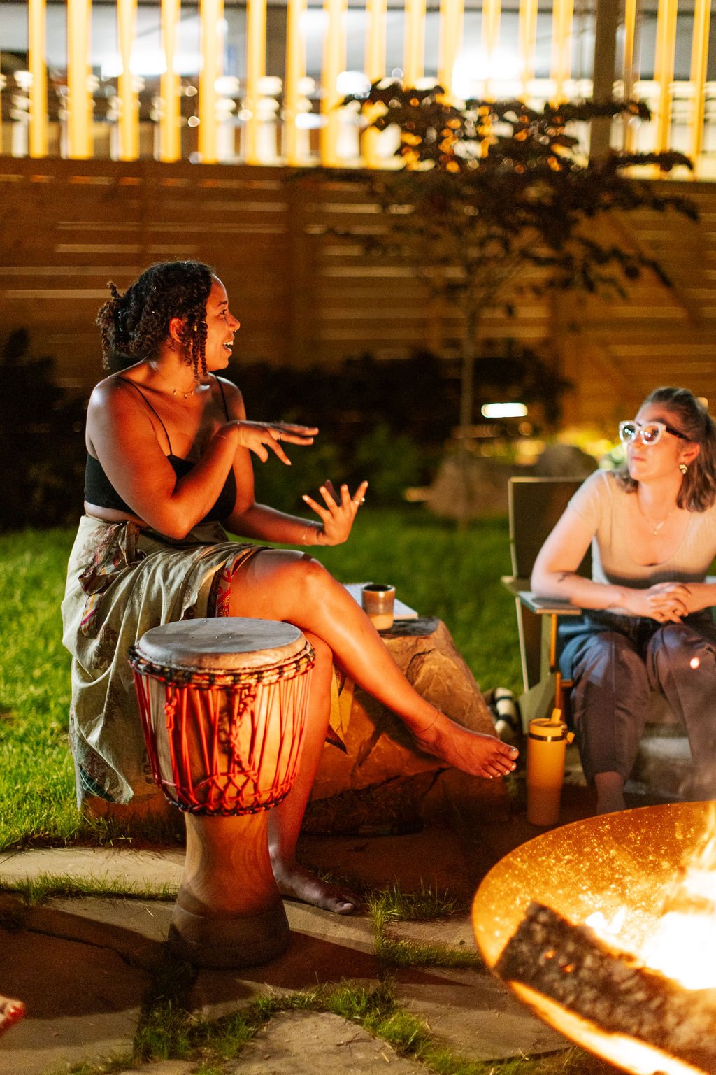Two women sitting outdoors at night, one is speaking and the other listening, with a fire in front of them, surrounded by grass and a wooden fence.
